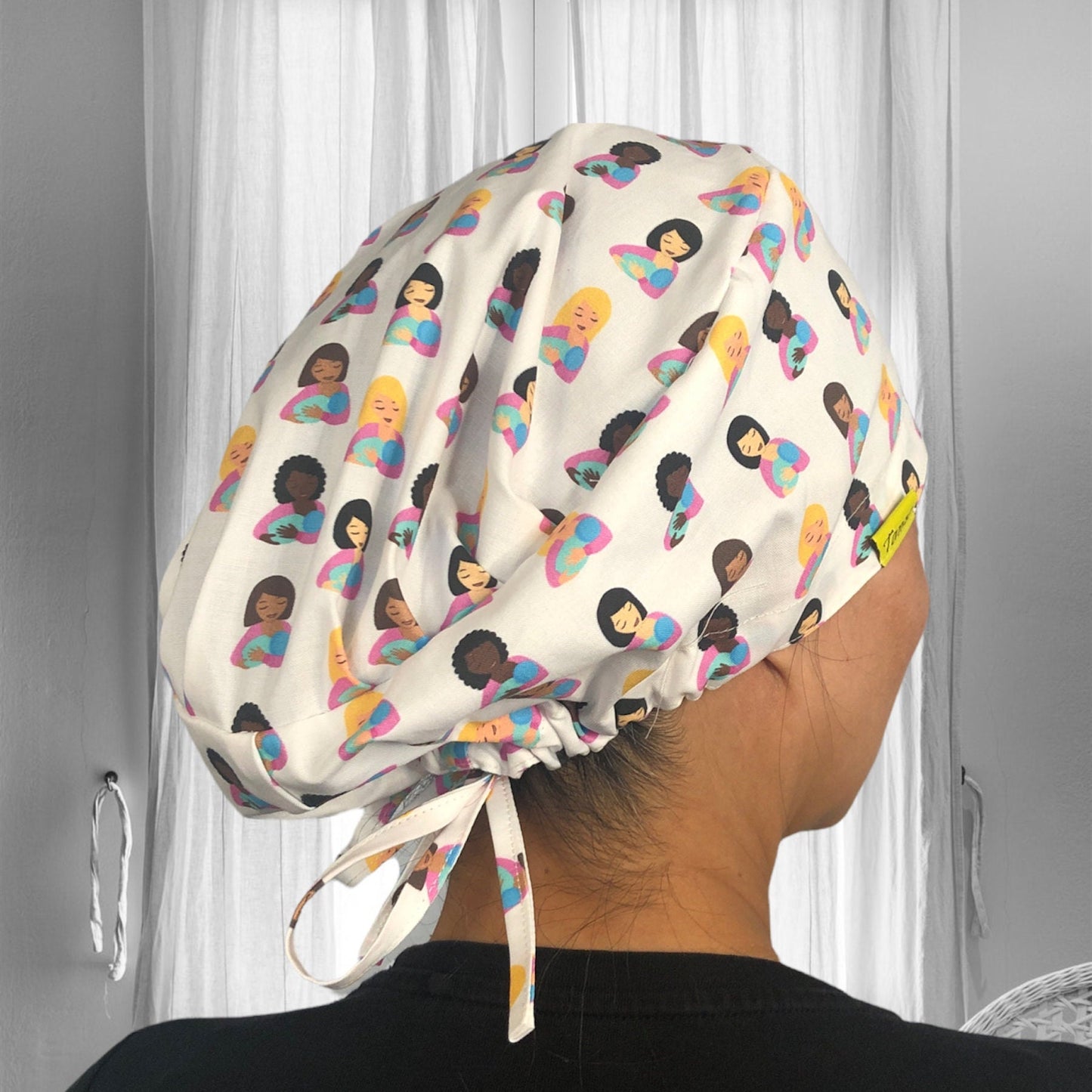 A soft, patterned scrub cap featuring diverse illustrations of women breastfeeding her baby against a white, textured background. The design includes various hair colors and skin tones, creating a vibrant and inclusive look.