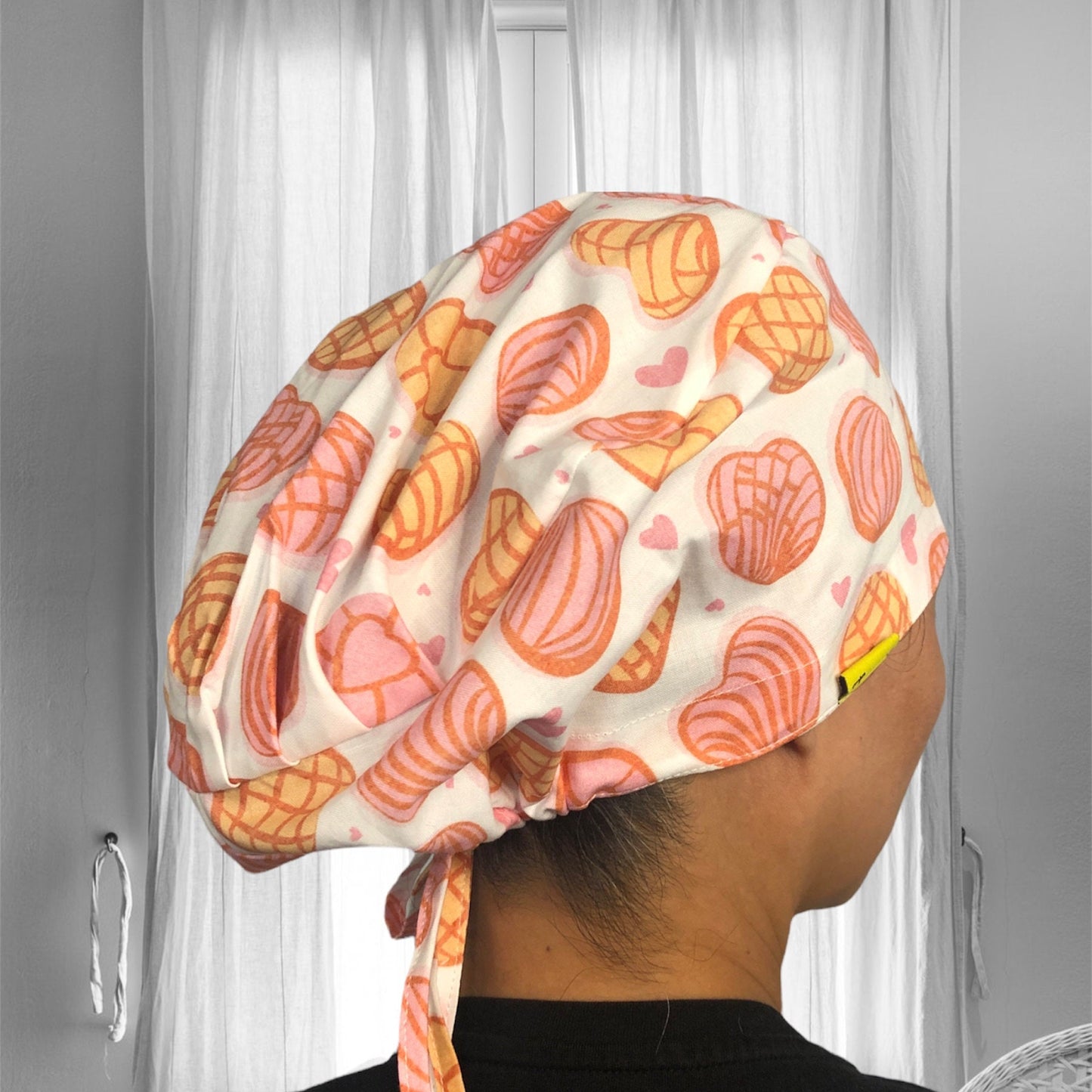 A person wearing a scrub cap with a playful design. The cap is adorned with a pattern featuring pink and orange conchas and hearts. It is tied at the back.