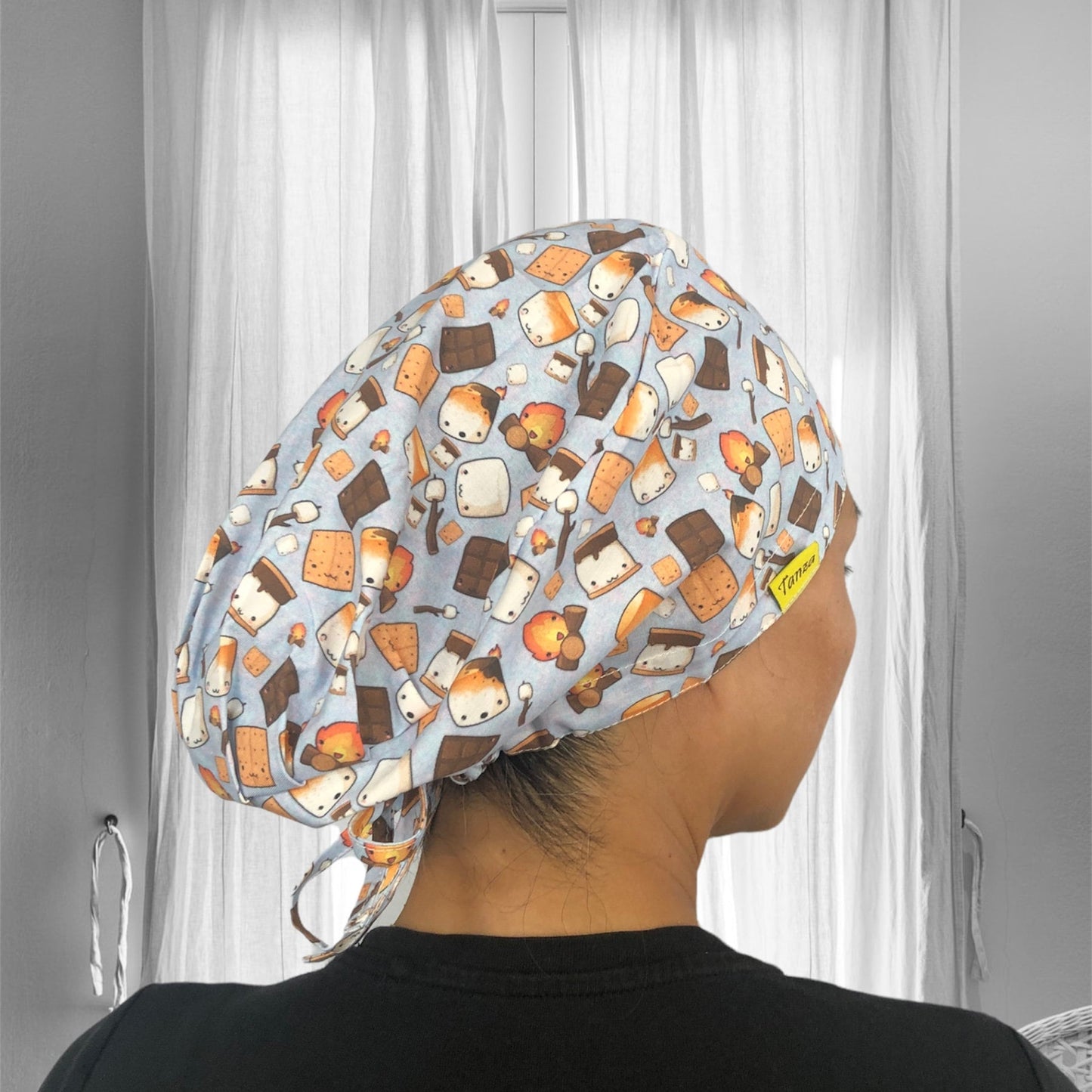 A person is wearing a colorful scrub cap featuring a playful pattern of cartoon s'mores, chocolate pieces, and marshmallows on a light blue background.