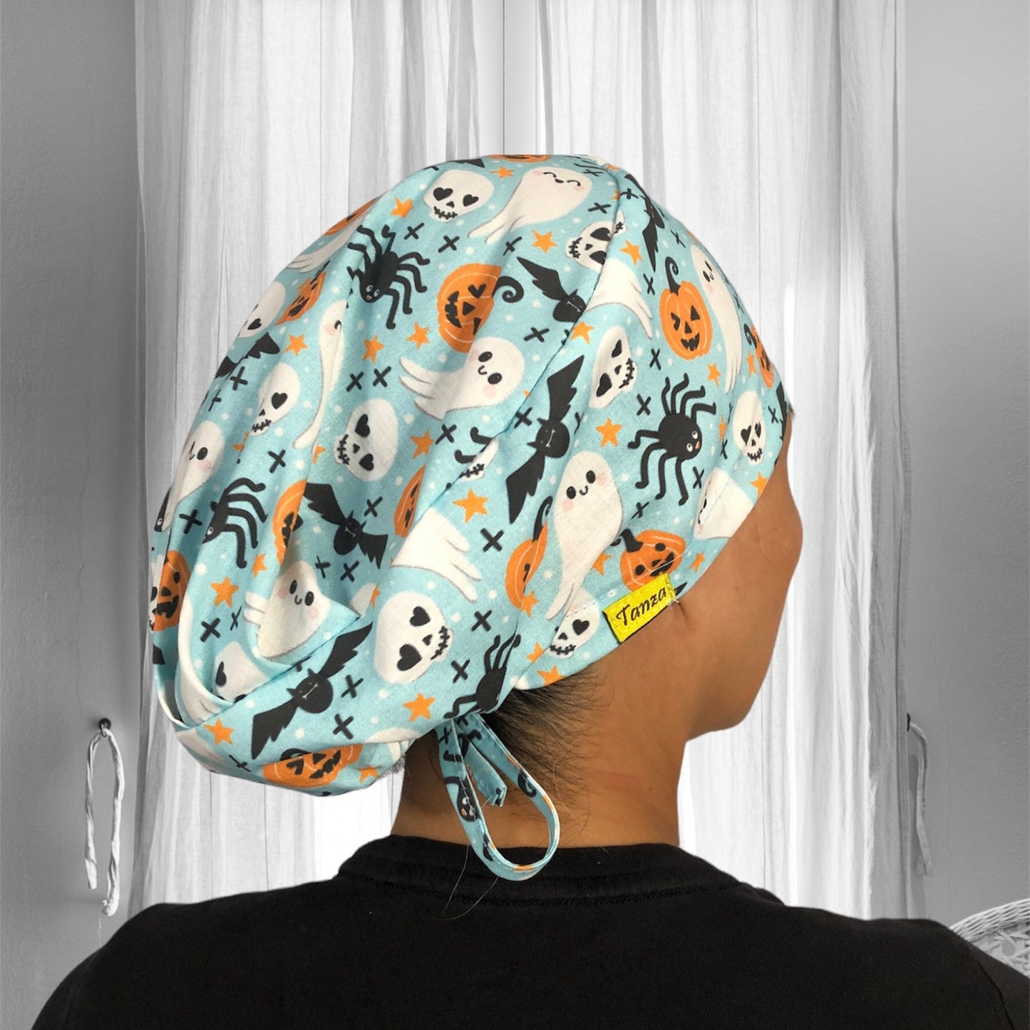 A person is wearing a colorful scrub cap with a Halloween theme. The fabric is adorned with playful designs of ghosts, skulls, bats, spiders, and orange pumpkins, set against a light blue background.