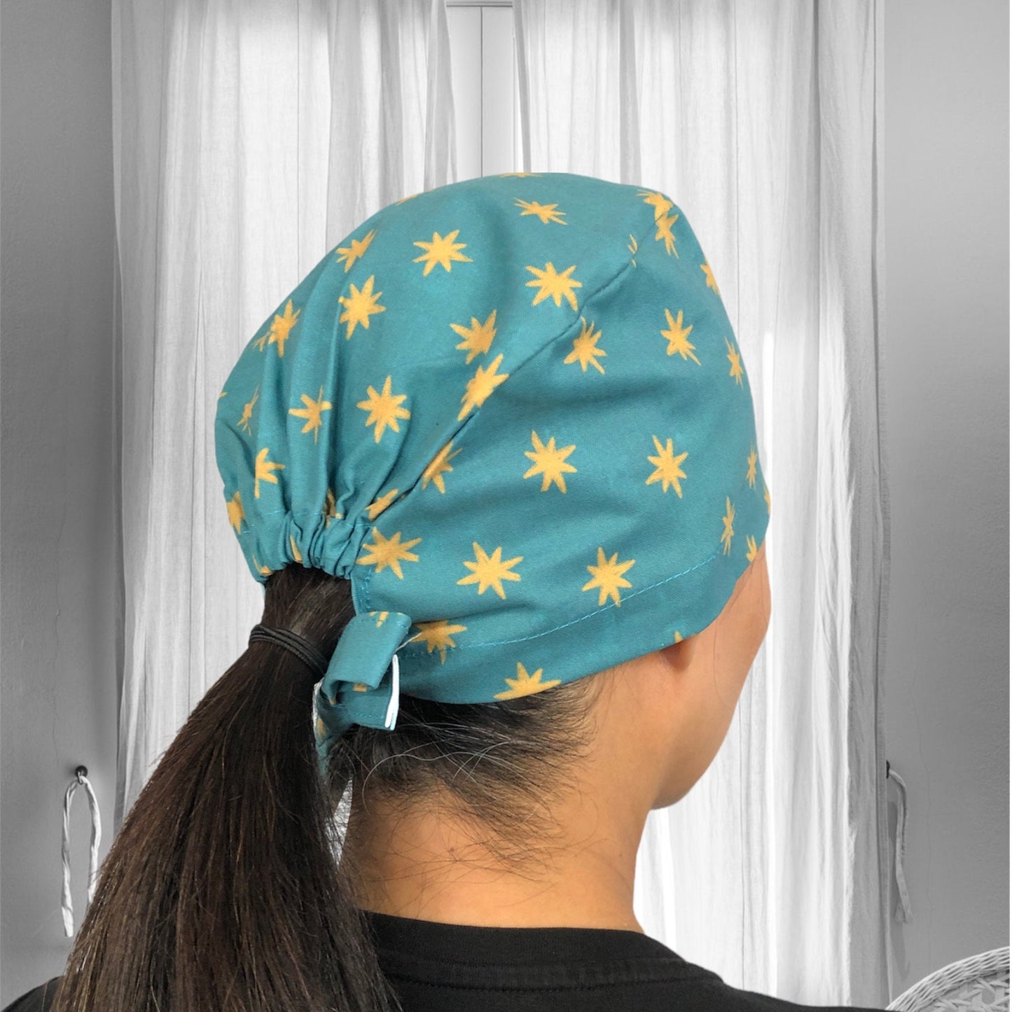 A person wearing a teal scrub cap with yellow star patterns, styled with a ponytail extending from the back.