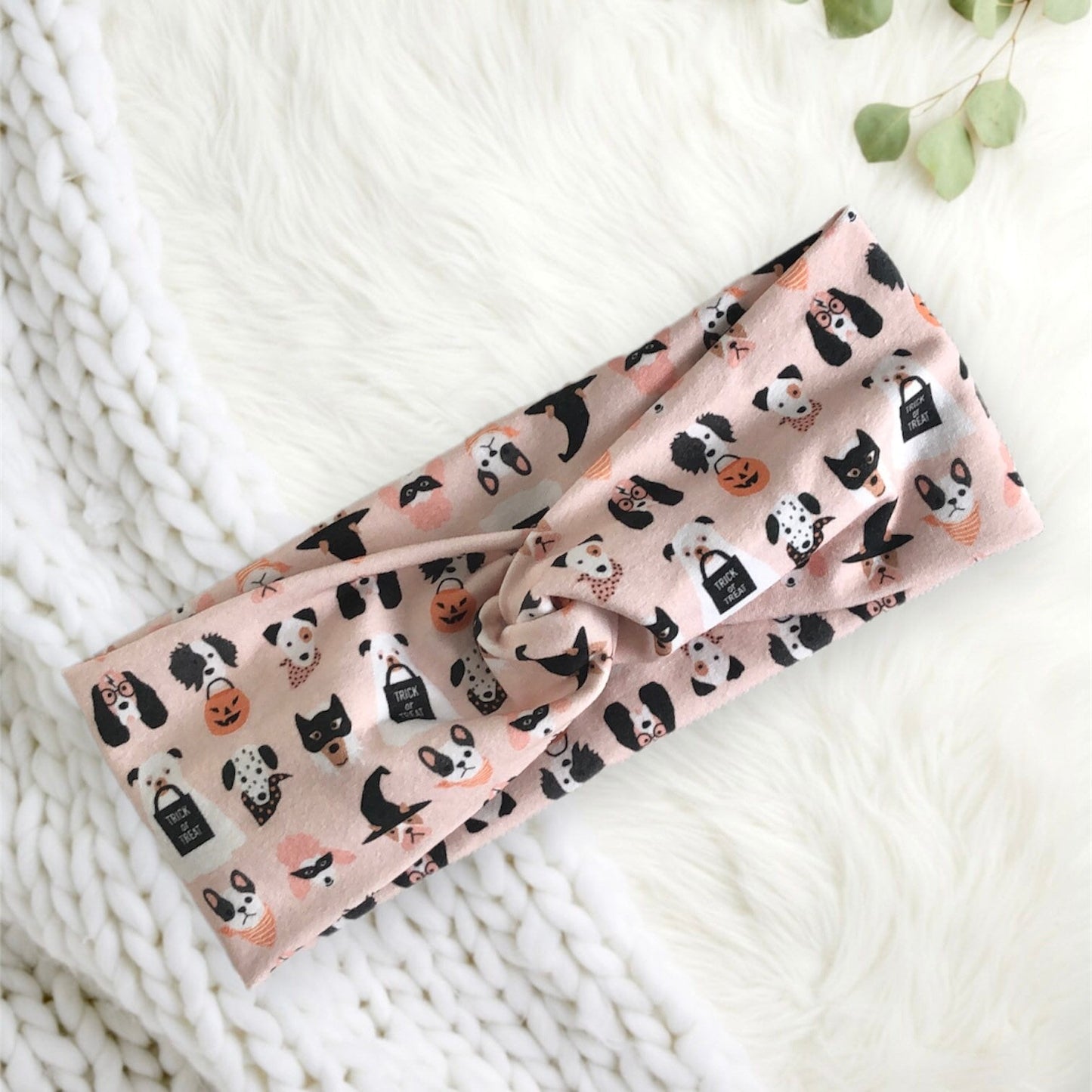 A soft, pastel pink headband adorned with playful Halloween-themed patterns, featuring cute illustrations of cats, dogs, pumpkins, and "trick or treat" signs. The headband is elegantly twisted at the center.