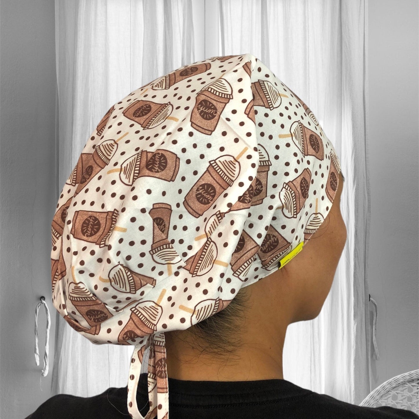 A person wearing a scrub cap adorned with a playful coffee theme pattern, featuring cups and coffee beans against a backdrop of white with brown polka dots.
