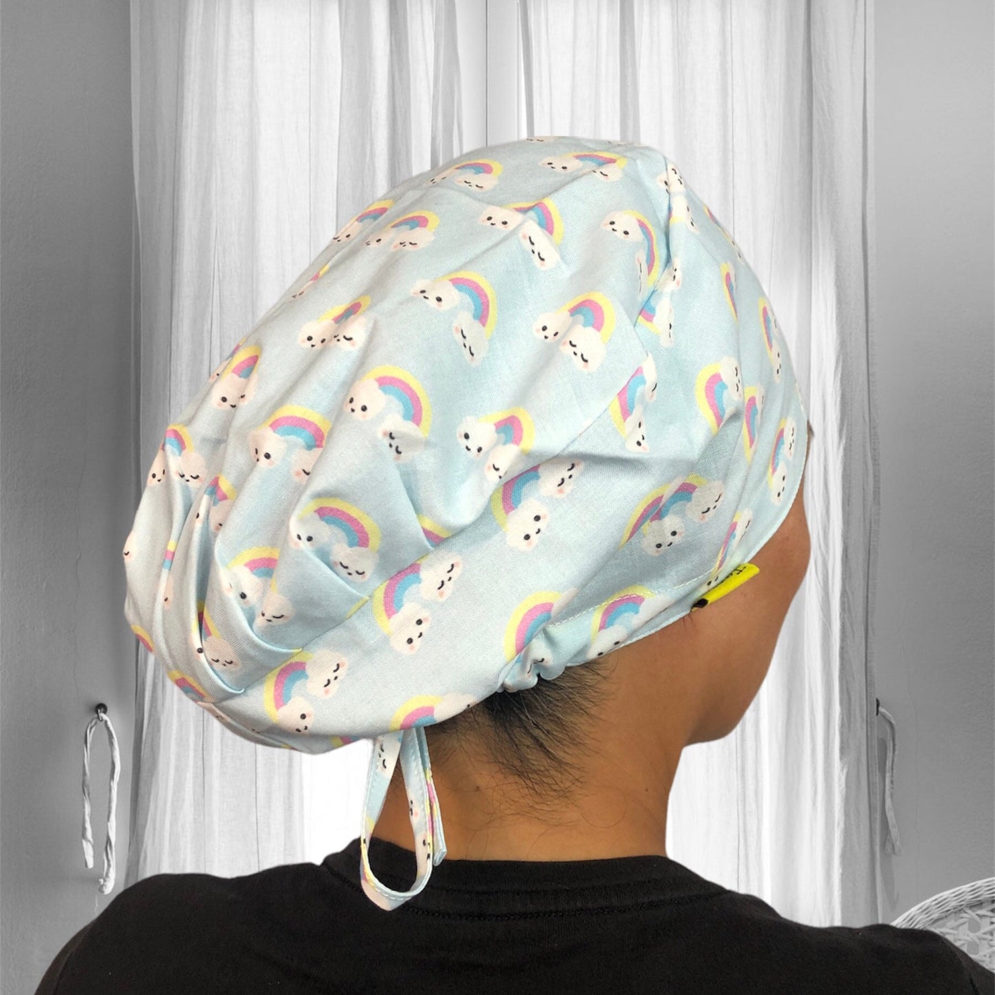 A person wearing a light blue scrub cap adorned with a cute pattern featuring smiling clouds and rainbows.