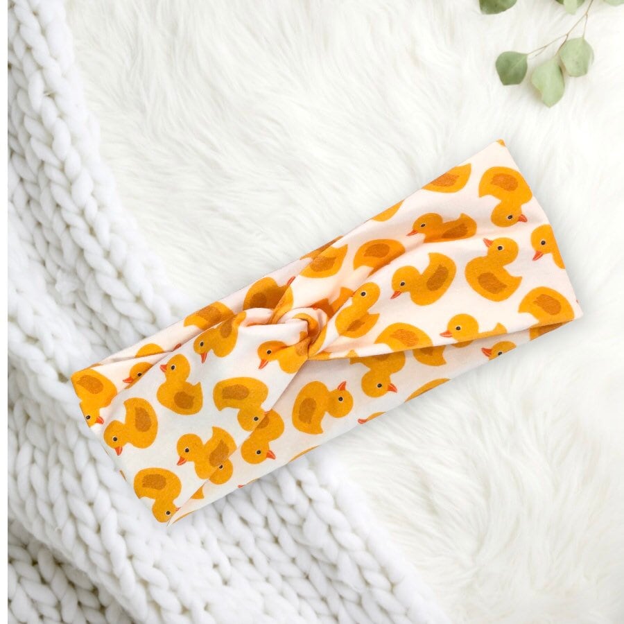 A soft, knotted headband adorned with a playful pattern of yellow rubber ducks on a white background.