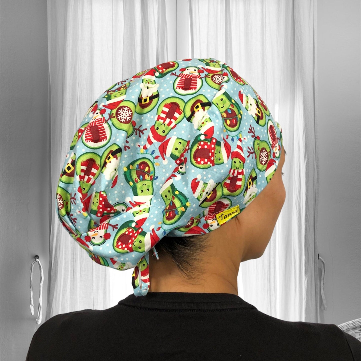 A colorful scrub cap adorned with festive holiday-themed prints. The cap features illustrations of snowmen, candy canes, and Christmas stockings set against a light blue background.