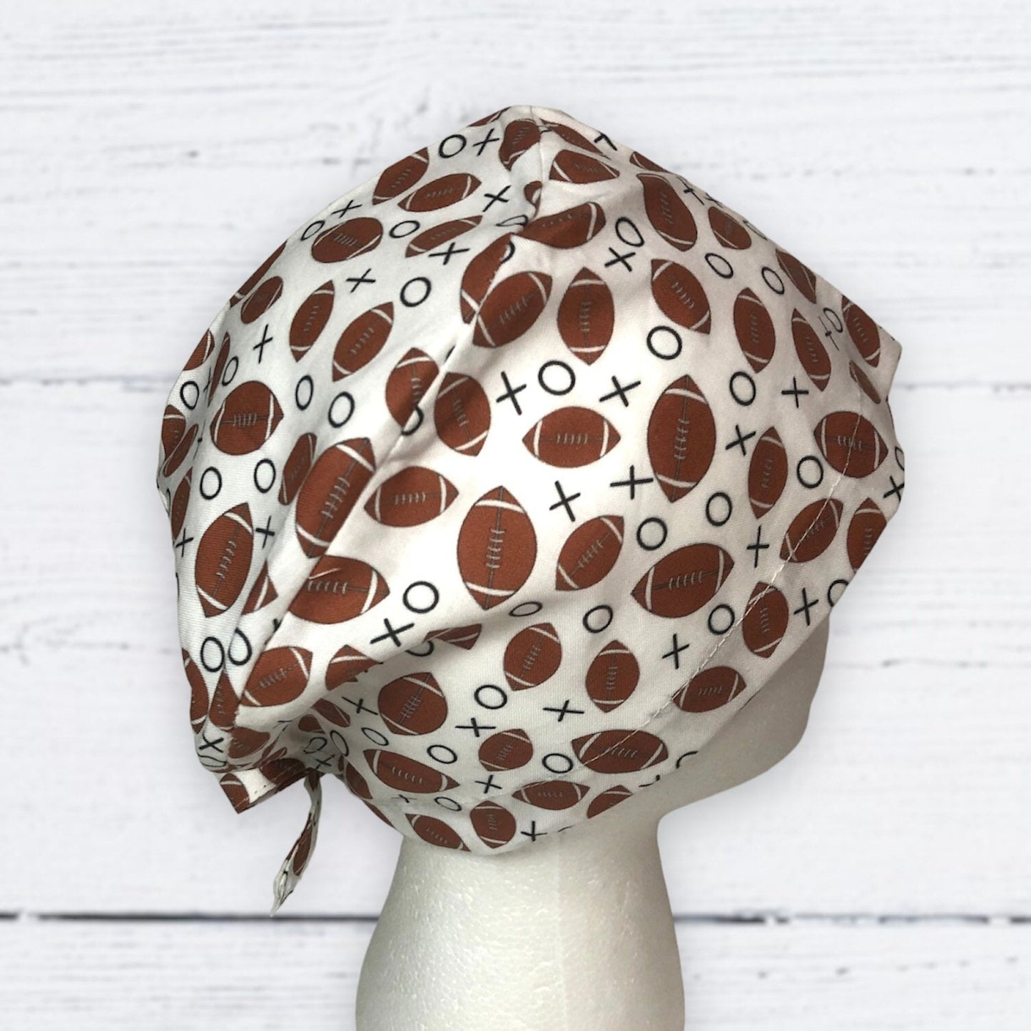 A white scrub cap with a fun and sporty design featuring brown American footballs and black X and O symbols scattered across the fabric.
