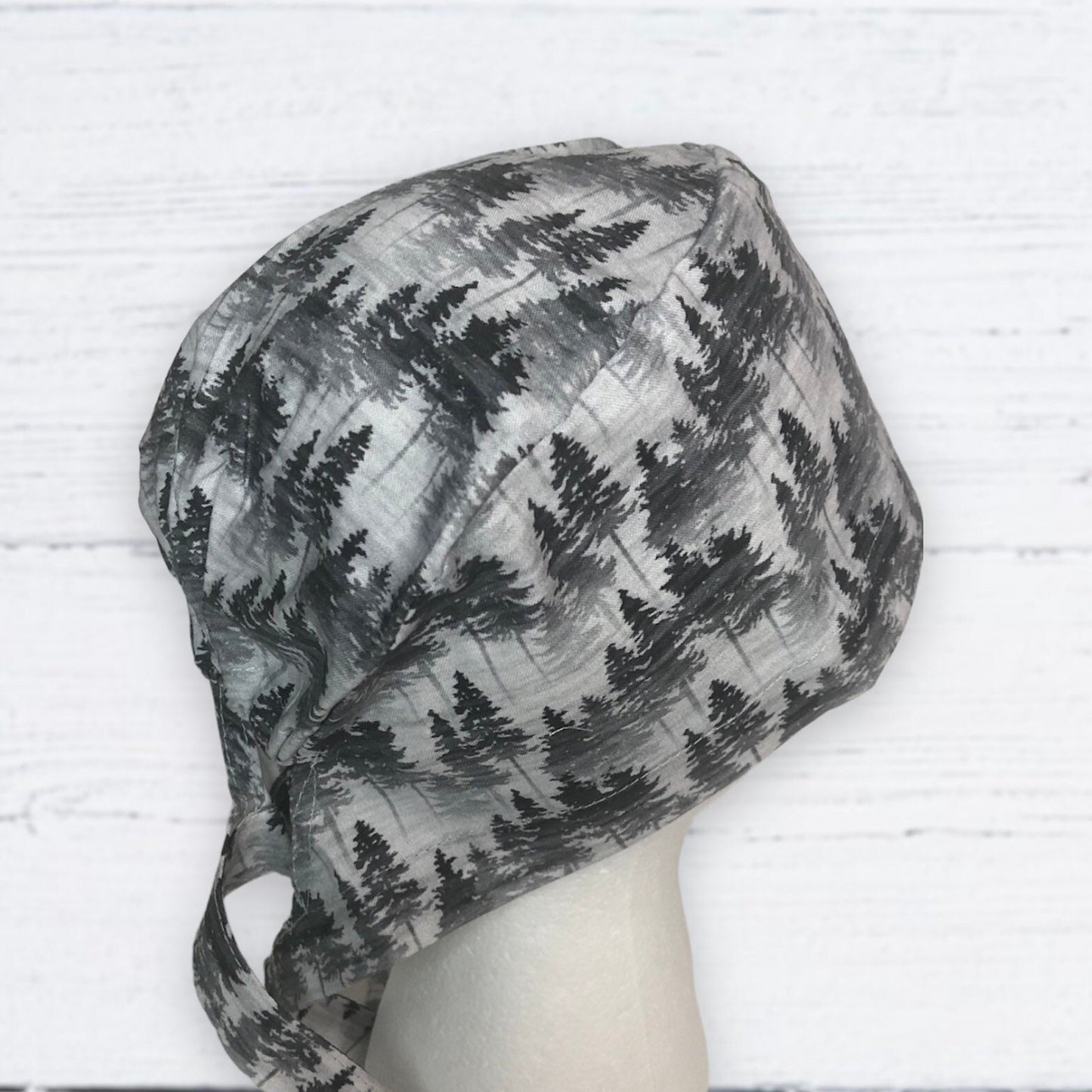 A grey fabric scrub cap featuring a pattern of dark green pine trees, set against a light grey background.