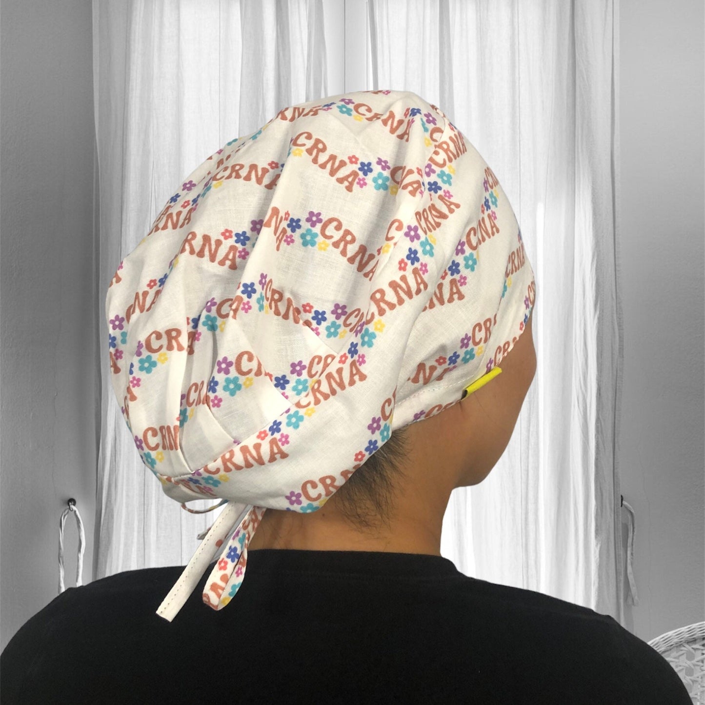 A person wearing a scrub cap with a print featuring the letters "CRNA" and colorful flowers.