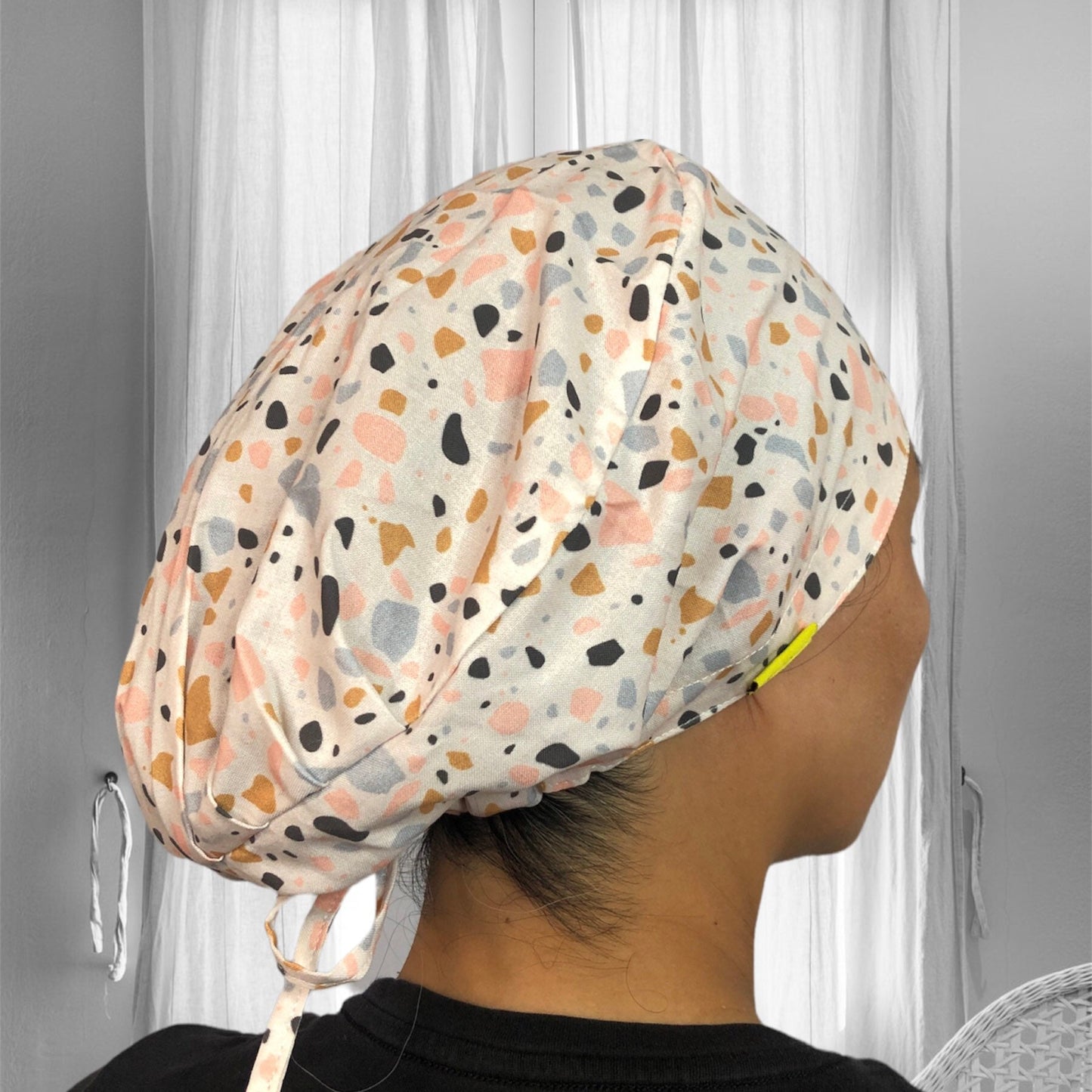 A person wearing a stylish scrub cap with a colorful terrazzo pattern. The cap features pastel shades of pink, blue, orange, and black speckles on a light background.