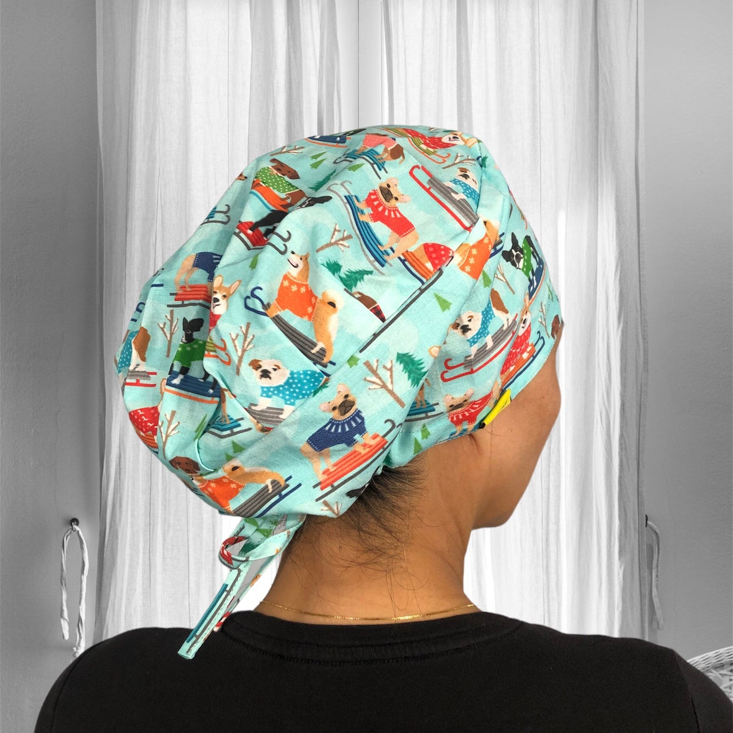 A person is wearing a colorful scrub cap featuring playful illustrations of dogs in various outfits and poses. The fabric is light blue with images of dogs sledding, some wearing sweaters.