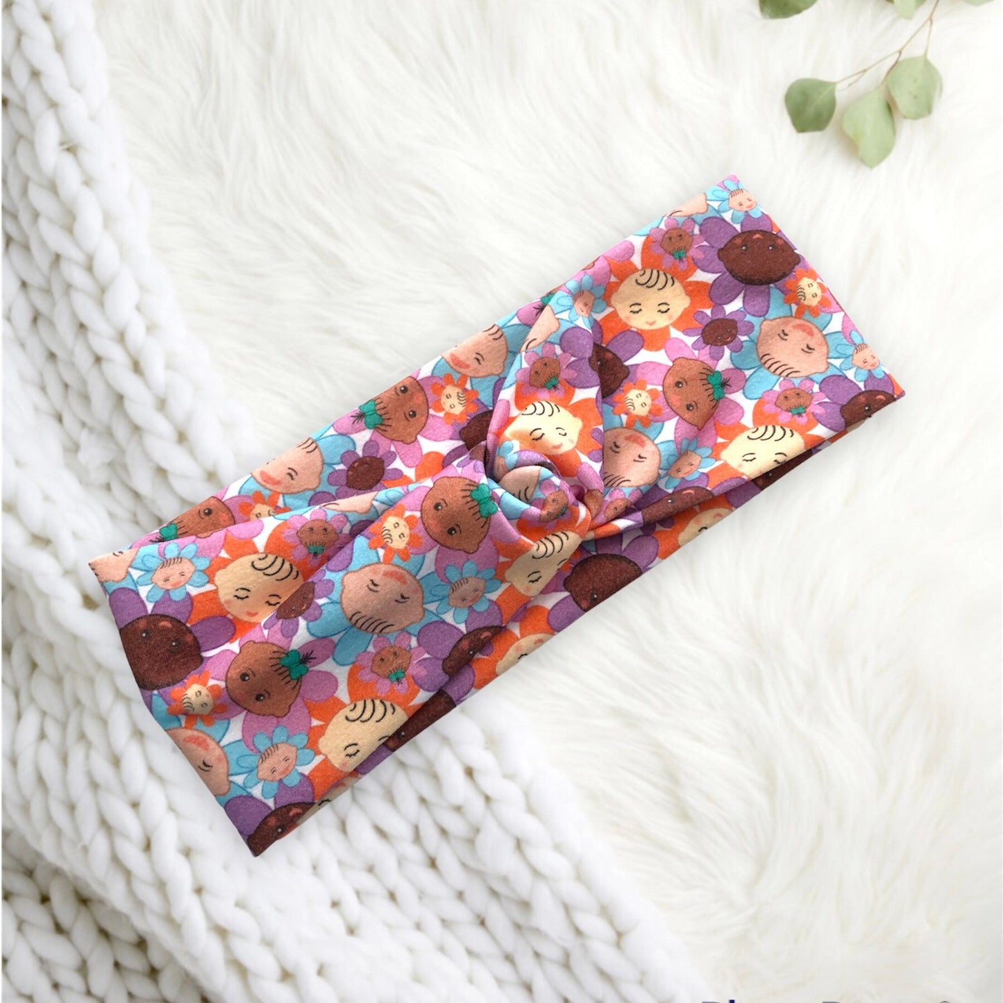 A colorful headband featuring a playful pattern of smiling baby faces and flowers in shades of orange, purple, and blue, resting on a soft, fluffy white background. The headband is neatly folded with a subtle twist in the center.