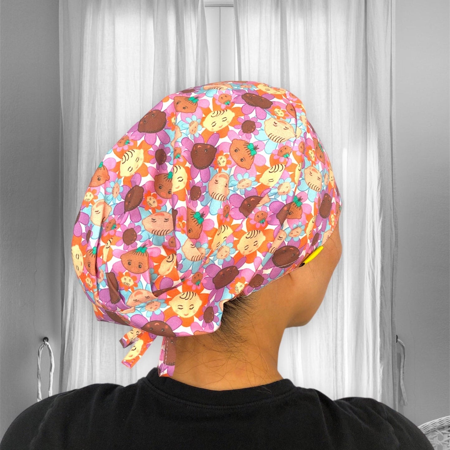 A colorful scrub cap featuring a fun and playful pattern of cheerful cartoon baby faces and flowers.
