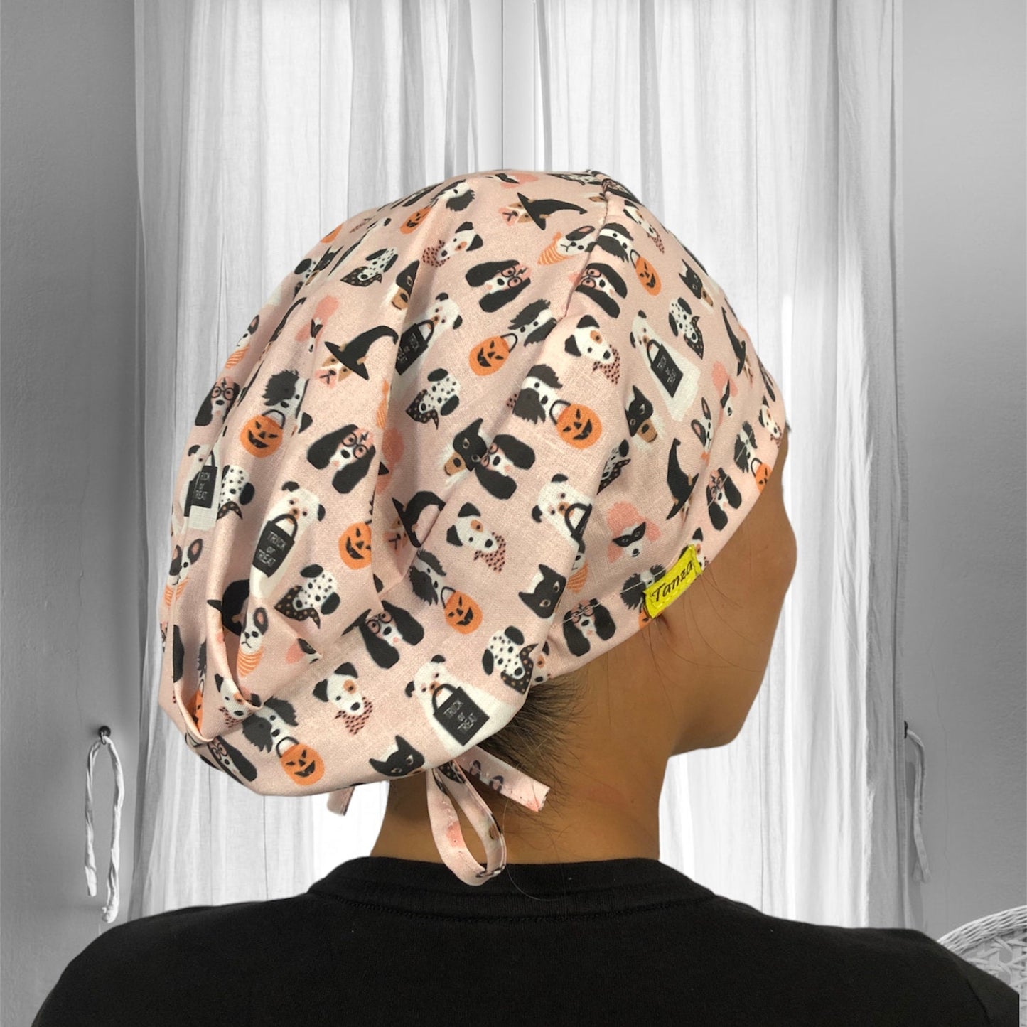 A soft, pastel pink scrub cap adorned with playful Halloween-themed patterns, featuring cute illustrations of cats, dogs, pumpkins, and "trick or treat" signs.