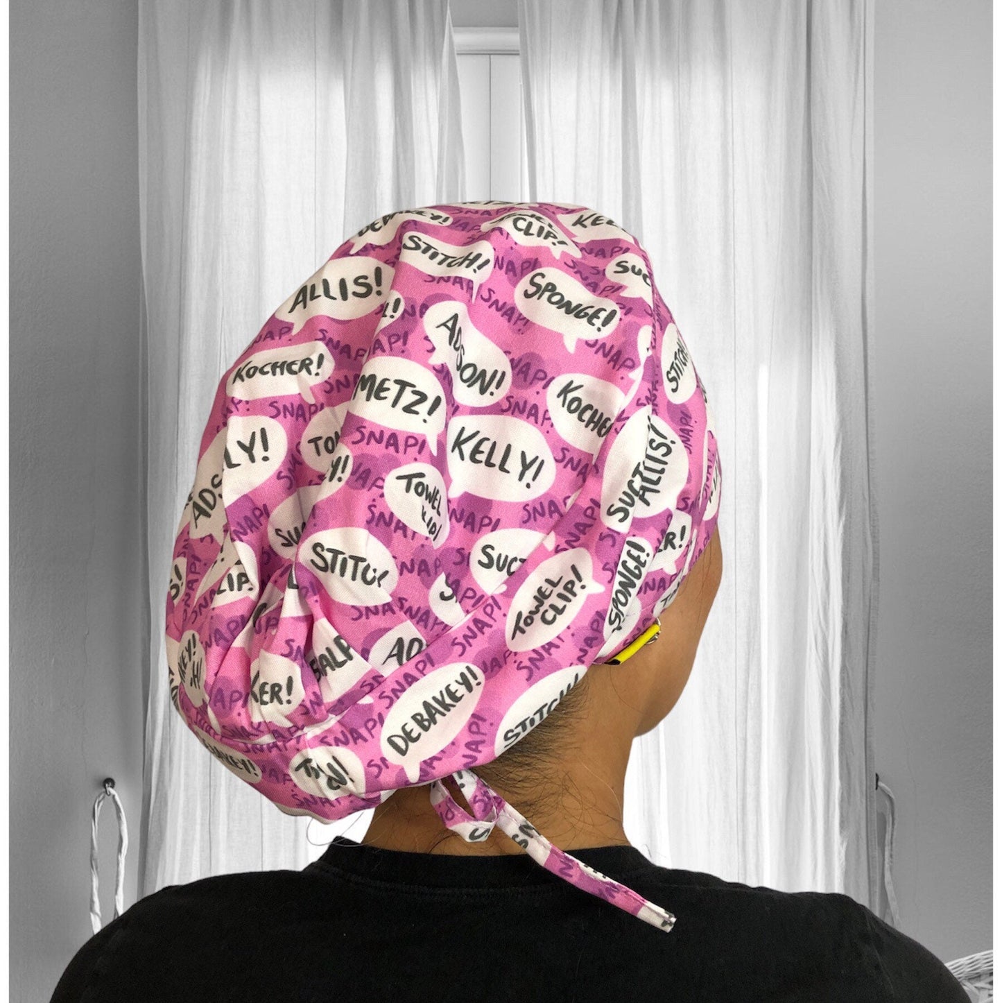 A pink scrub cap with a pattern of white speech bubbles, each containing medical and surgical terms such as "Sponge!", "Snap!", "Kocher!", "Debakey!", and more.