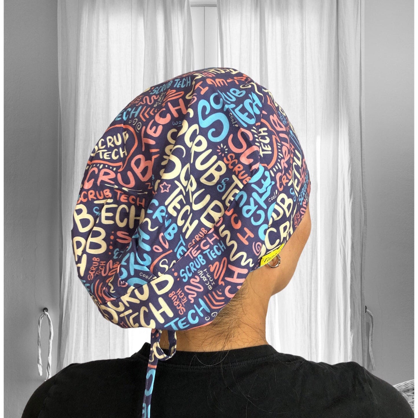 A colorful scrub cap with a vibrant pattern featuring the words "Scrub Tech" in various fonts and sizes. It highlights the design that includes shades of blue, orange, yellow, and pink.