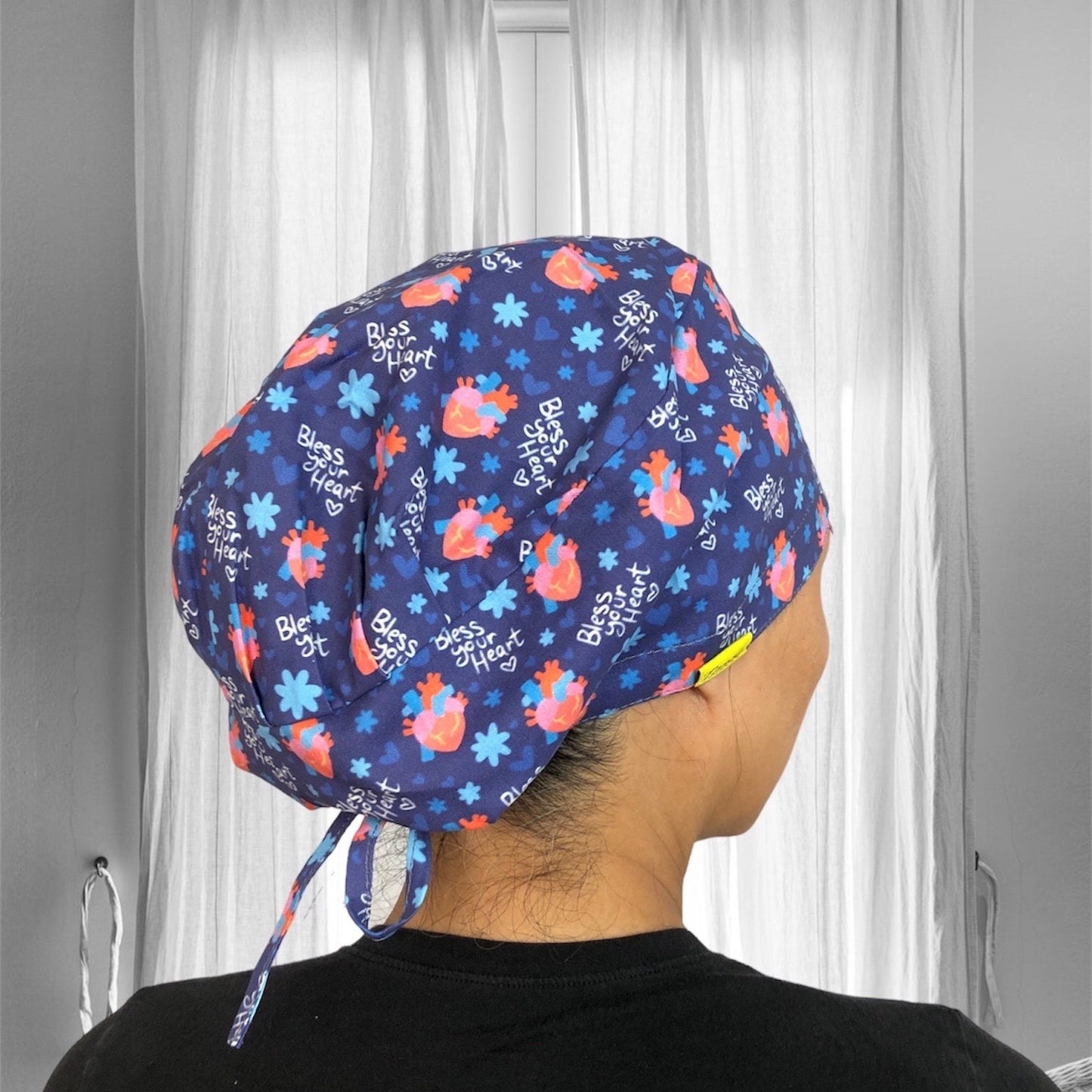 A blue scrub cap adorned with a pattern of hearts and flowers. The cap features the phrase "Bless Your Heart" printed repeatedly across the fabric.