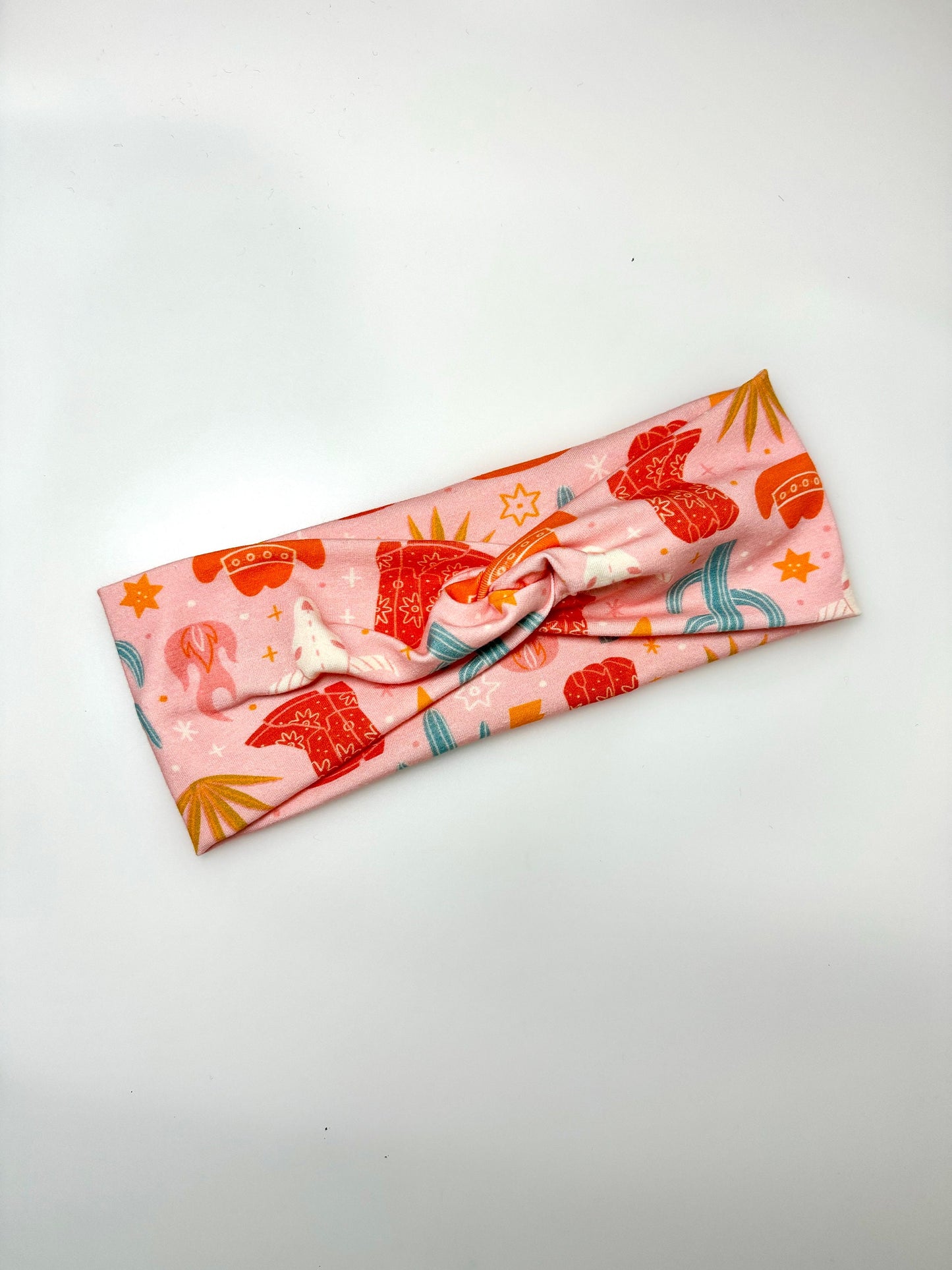 A pink headband featuring a playful pattern with red and orange motifs, including stars, cacti, and abstract shapes. The fabric is twisted into a knot at the center.