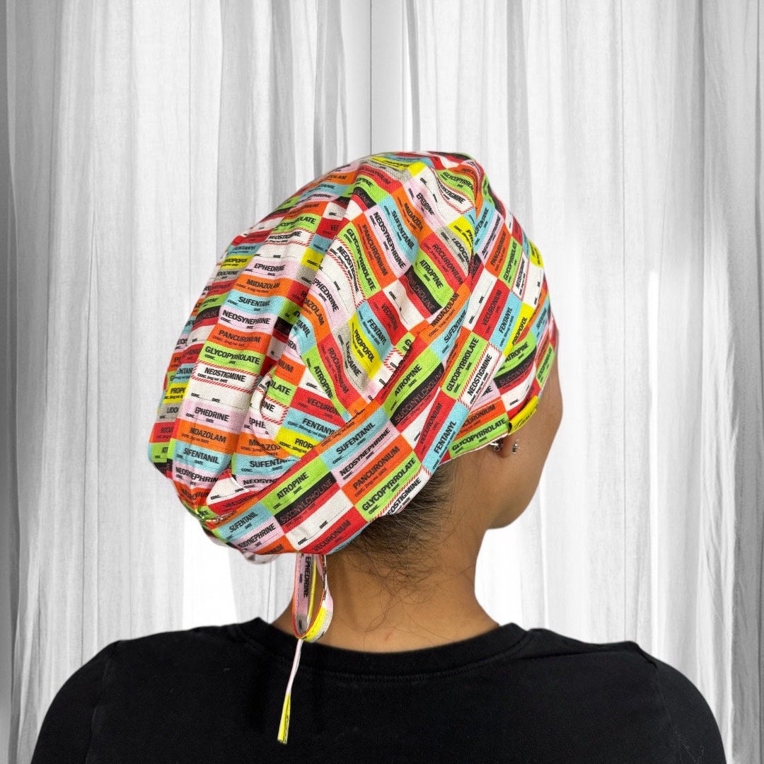 A person wearing a colorful scrub cap with a pattern of various medication names. The cap features a variety of vibrant colors including red, green, blue, and yellow, with each section labeled with a different drug name.