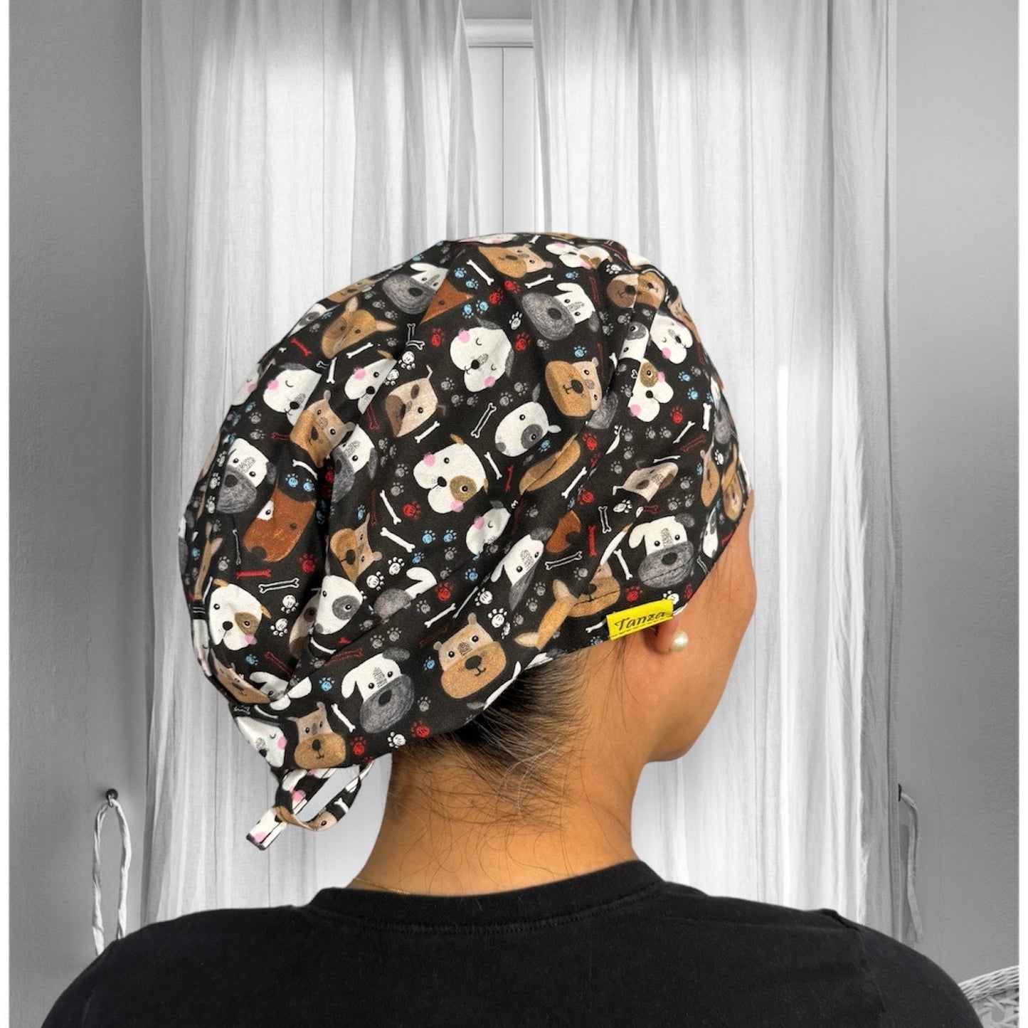 A person wearing a scrub cap featuring a playful dog pattern. The cap is adorned with various cartoon-style dog faces in different colors and expressions, set against a dark background. The fabric is decorated with small bones and paw prints, adding to the whimsical theme.