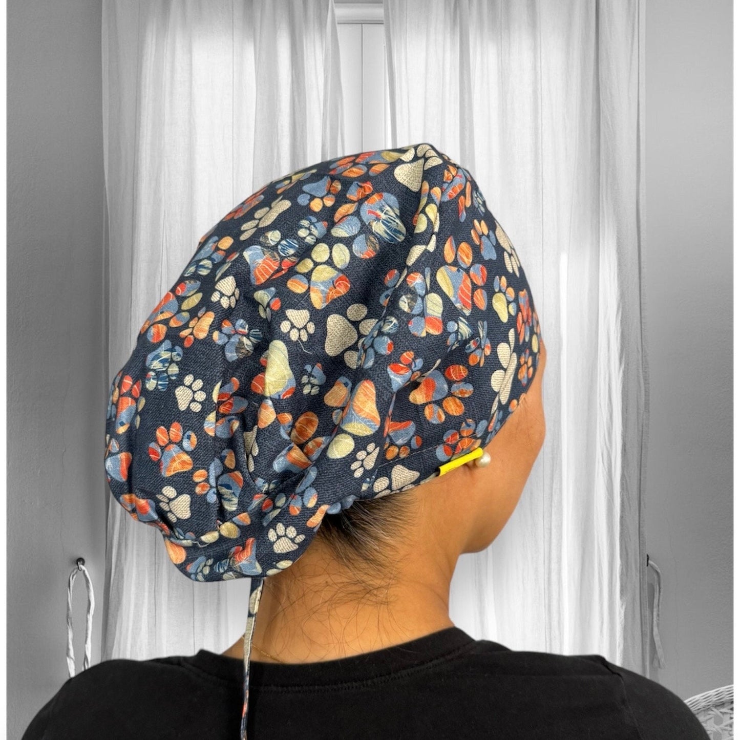 A person wearing a scrub cap featuring a colorful paw print pattern. The cap has a dark background with paw prints in shades of orange, cream, and blue.