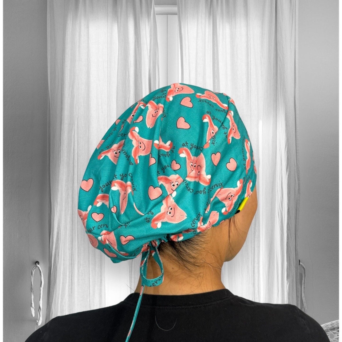 The cap features a teal background with cartoon illustrations of smiling cervixes surrounded by pink hearts. The text "at your cervix" is playfully repeated across the fabric.