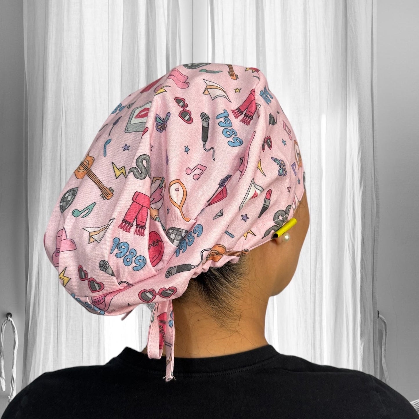 A stylish pink scrub cap adorned with various playful illustrations including lipstick, a snake, guitar, and a heart. The design features vibrant colors and elements reminiscent of retro aesthetics.