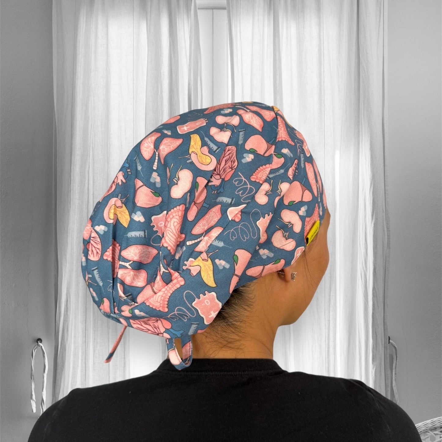 A person wearing a scrub cap with a playful pattern. The cap features various medical-themed illustrations in pink and yellow on a blue background.