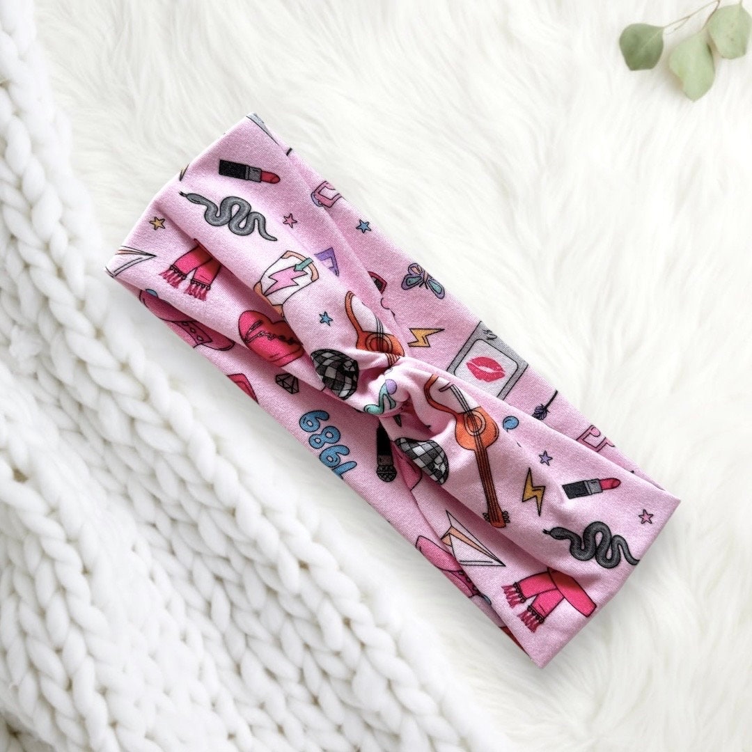 A stylish pink headband adorned with various playful illustrations including lipstick, a snake, guitar, and a heart. The design features vibrant colors and elements reminiscent of retro aesthetics.