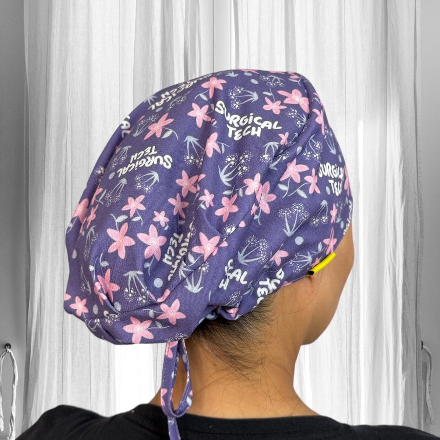 A person wearing a scrub cap with a colorful design. The cap is predominantly purple with light pink flowers and white text that reads "Surgical Tech."