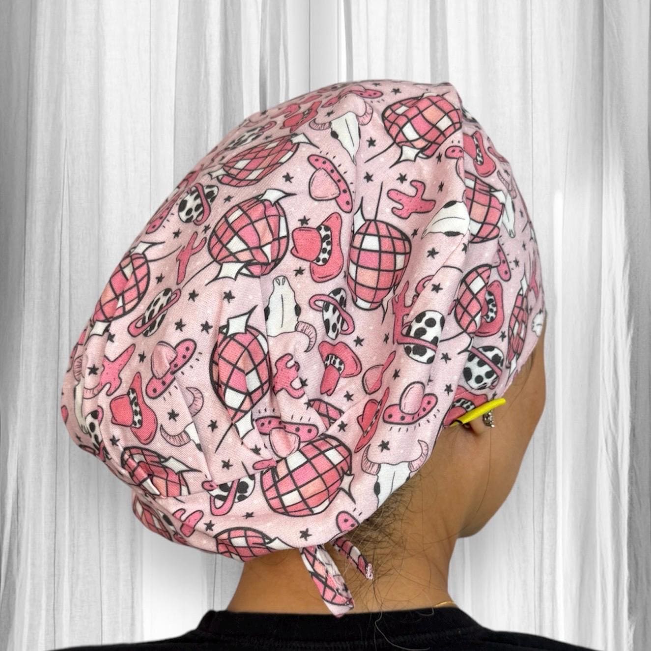 A scrub cap adorned with a playful design. The fabric is predominantly pink with various western-themed patterns, including cowboy hats, boots, and bandanas, depicted in shades of red, black, and white.