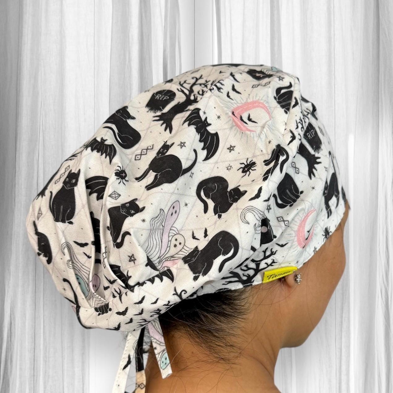 A scrub cap, featuring a Halloween-themed design with black cats, bats, ghosts, and other spooky elements on a light background. The fabric's print includes whimsical illustrations such as crescent moons and stars, adding a playful touch to the eerie motif.