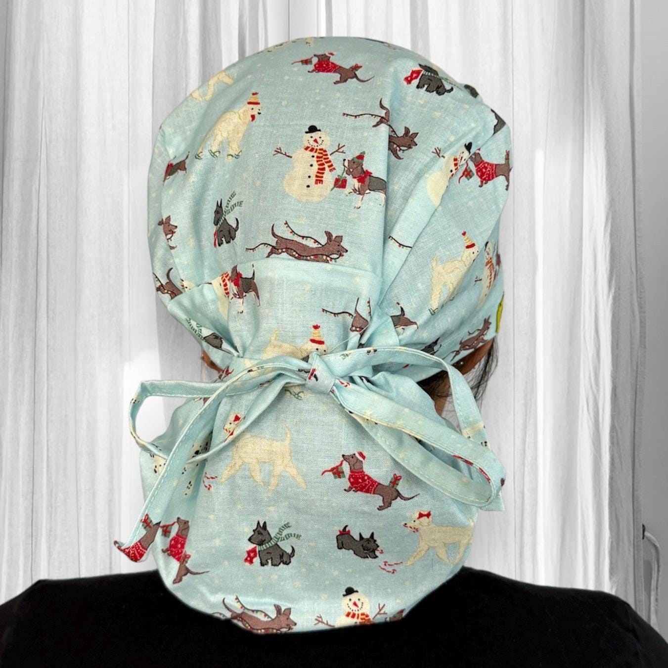A light blue fabric surgical cap adorned with whimsical winter-themed patterns. The design features snowmen, and dogs in festive attire, all scattered across the fabric.