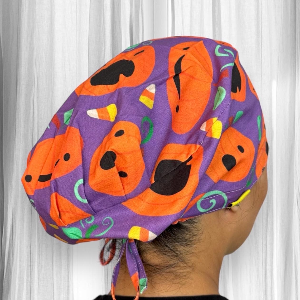A person wearing a vibrant scrub cap featuring a Halloween-themed design. The cap is adorned with orange pumpkin faces and candy corn motifs set against a purple background.