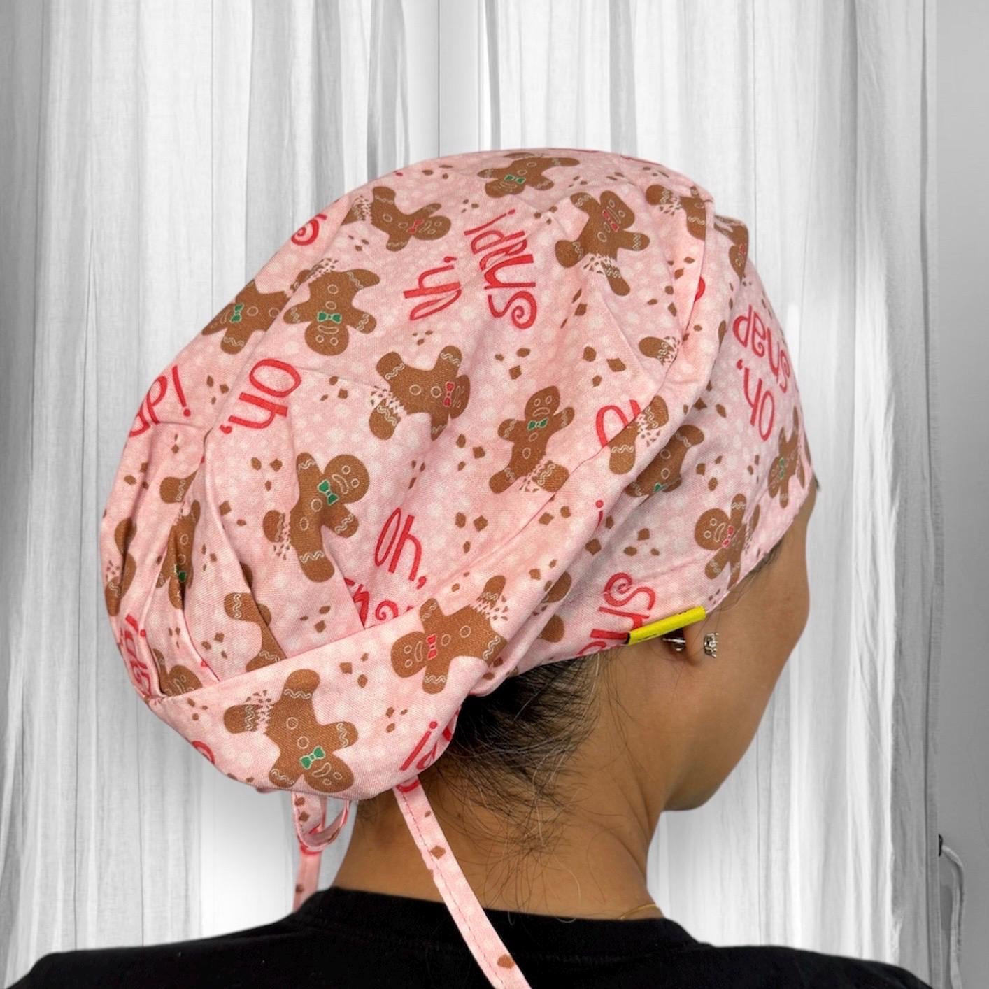 A stylish scrub cap featuring a playful pattern with gingerbread cookie characters and the text "oh snap" on a pink background.