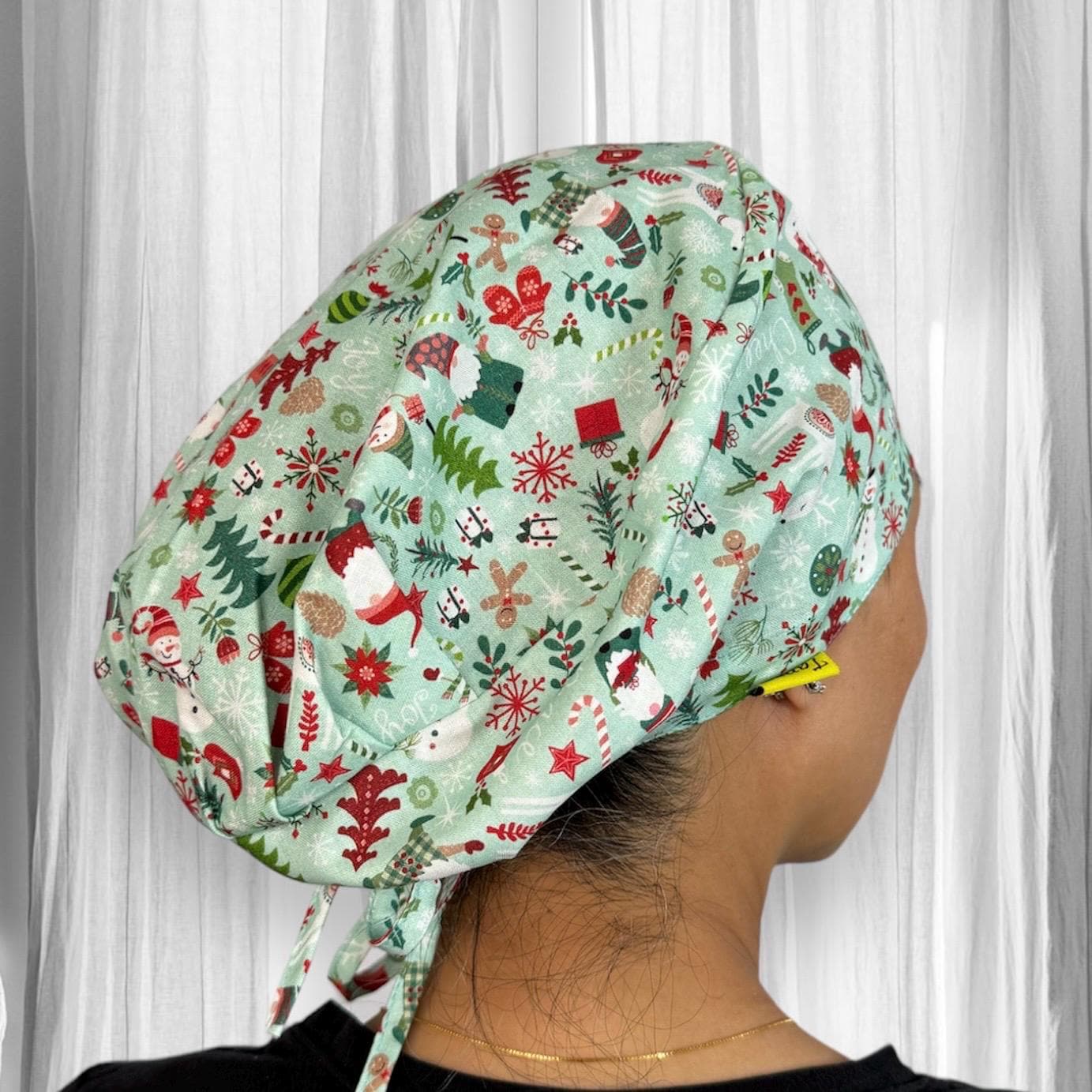 A person wearing a colorful scrub cap with a festive design. The cap features patterns of Christmas-themed elements like snowflakes, candy canes, holly, Christmas trees, snowmen, and reindeer on a light green background.