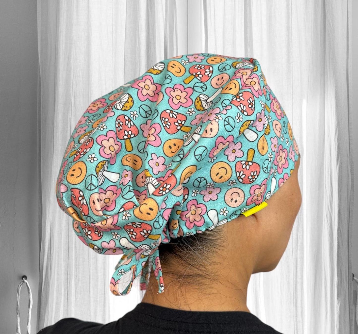 A person wearing a colorful scrub cap with a vibrant and playful pattern. The cap features a variety of designs, including peace signs, mushrooms, flowers, and smiley faces, all in shades of teal, pink, and orange.