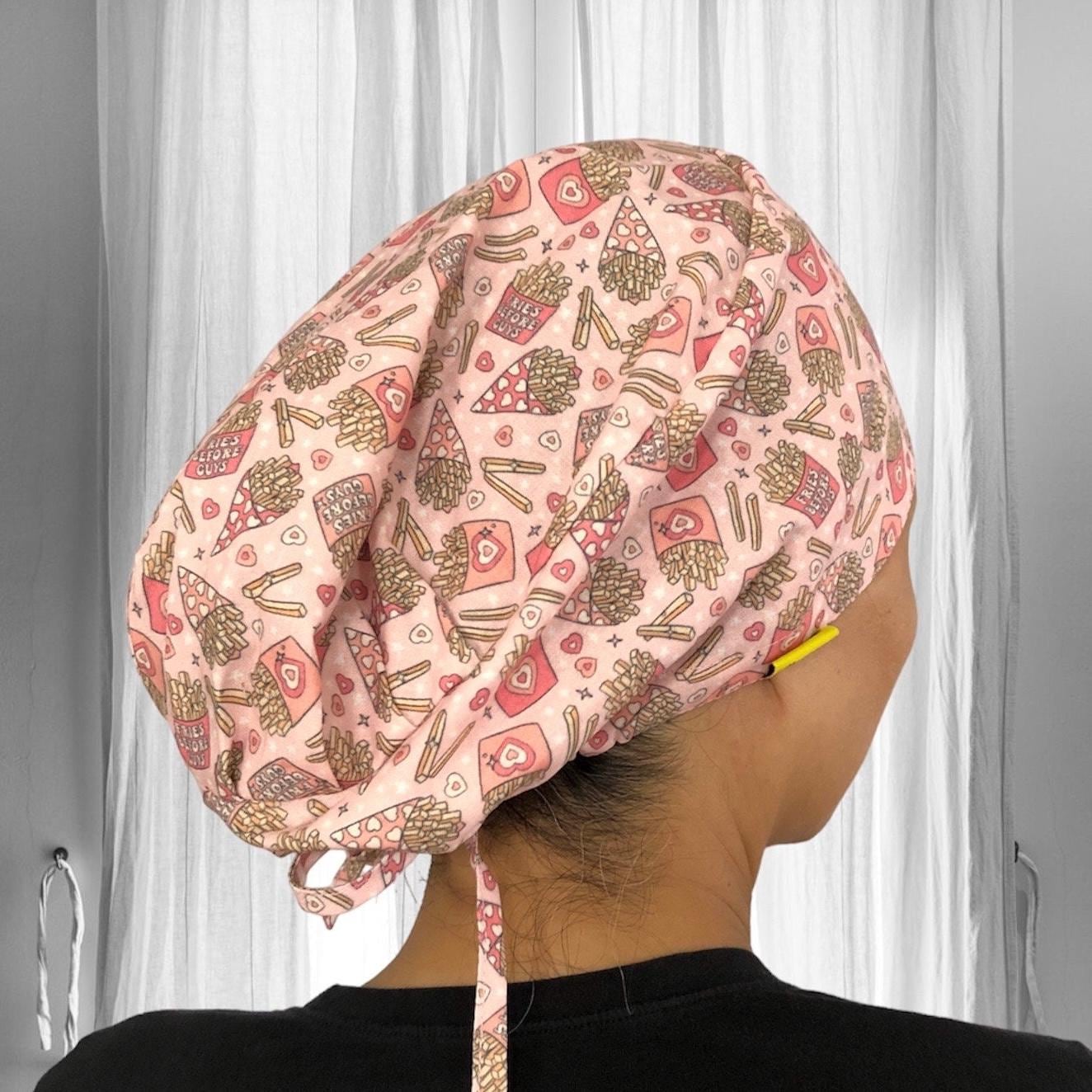 A person wearing a scrub cap with a playful pattern featuring pizza slices, French fries, and text bubbles with phrases like "Food Before Guys". The cap has a pink background with a variety of food-themed illustrations in red, brown, and yellow colors.