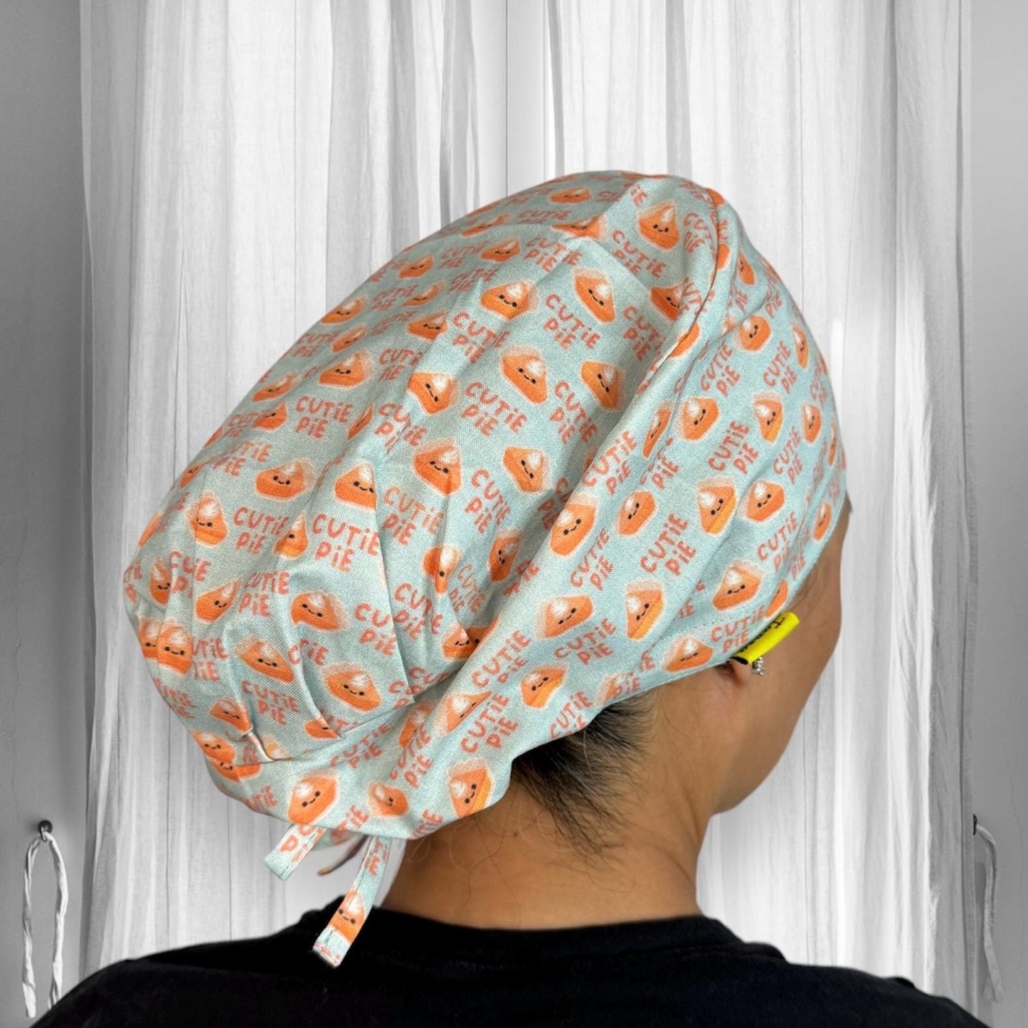 A person wearing a scrub cap with a playful design featuring orange pie graphics and the words "Cutie Pie" repeated across a light blue fabric.