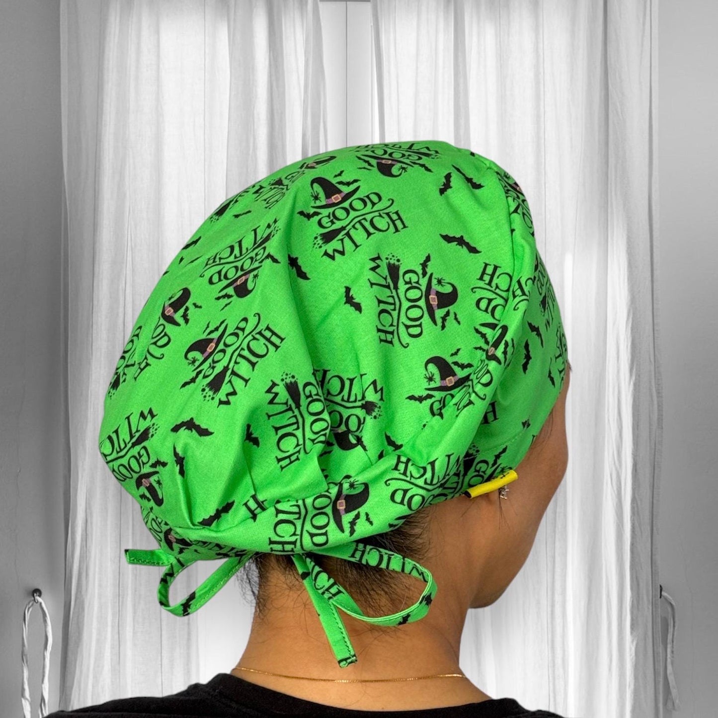 A person is wearing a vibrant green scrub cap featuring the playful phrase "Good Witch" alongside witch hats and bats.