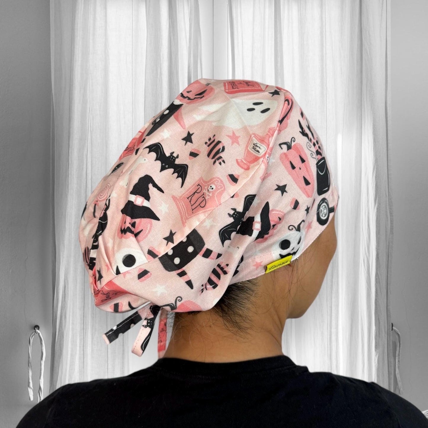 A person wearing a scrub cap with a playful Halloween-themed design. The cap features various festive prints including pumpkins, bats, ghosts, and witches' hats, all in shades of pink, black, and white.