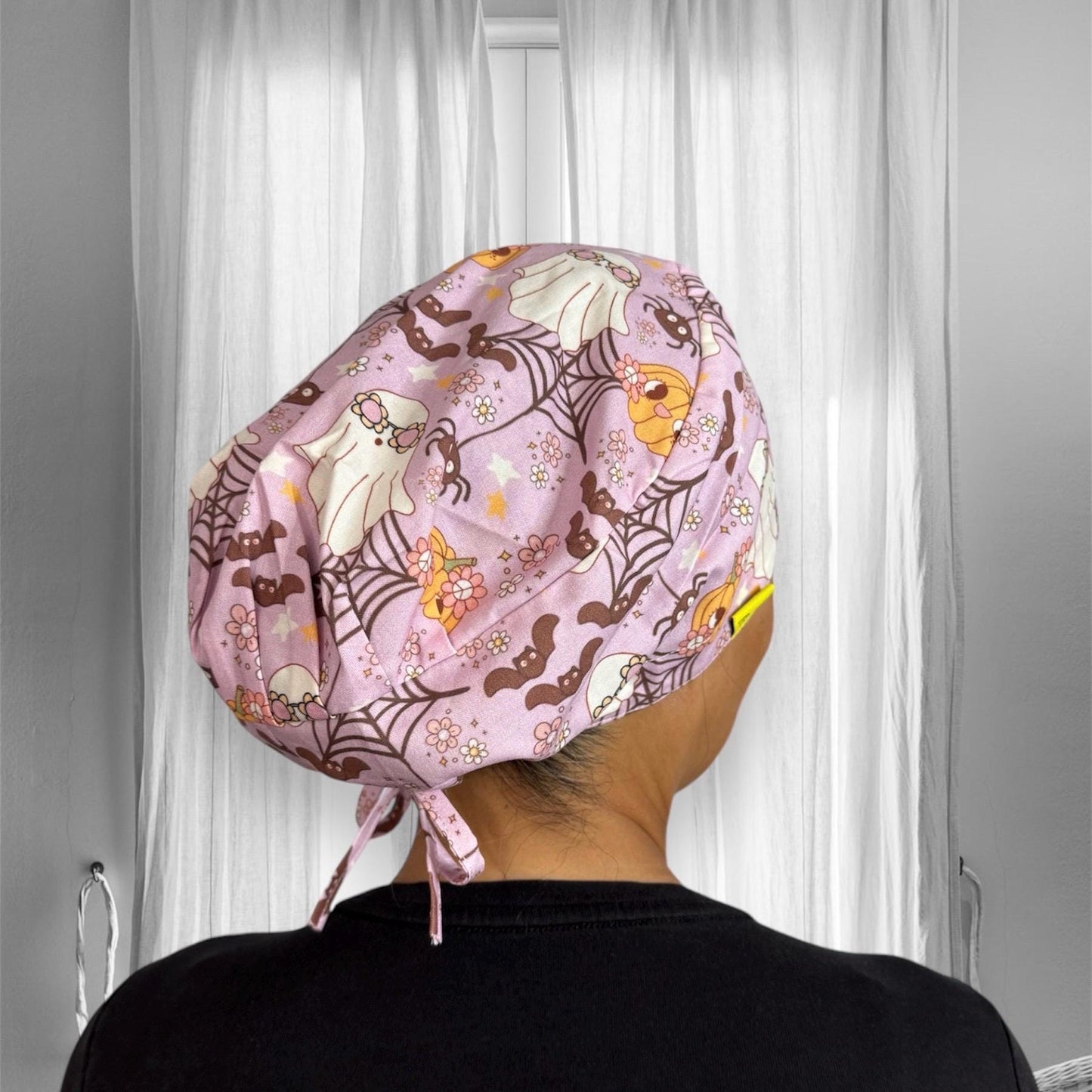 A person is wearing a pastel pink scrub cap adorned with a whimsical Halloween pattern. The design features cute ghosts, pumpkins, bats, and spider webs, all interspersed with small flowers.