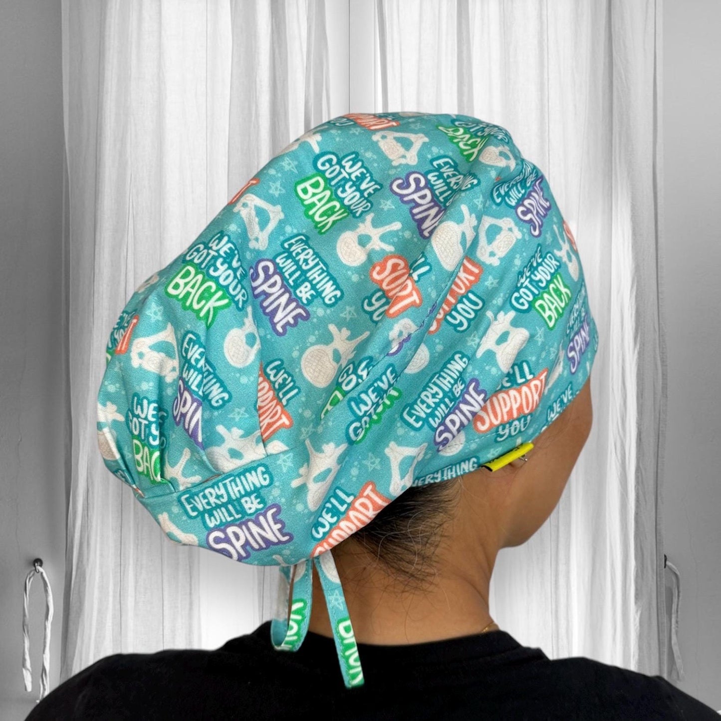 The scrub cap is adorned with playful orthopedic-themed phrases like "Everything will be spine," "We got your back," and simple spine illustrations.