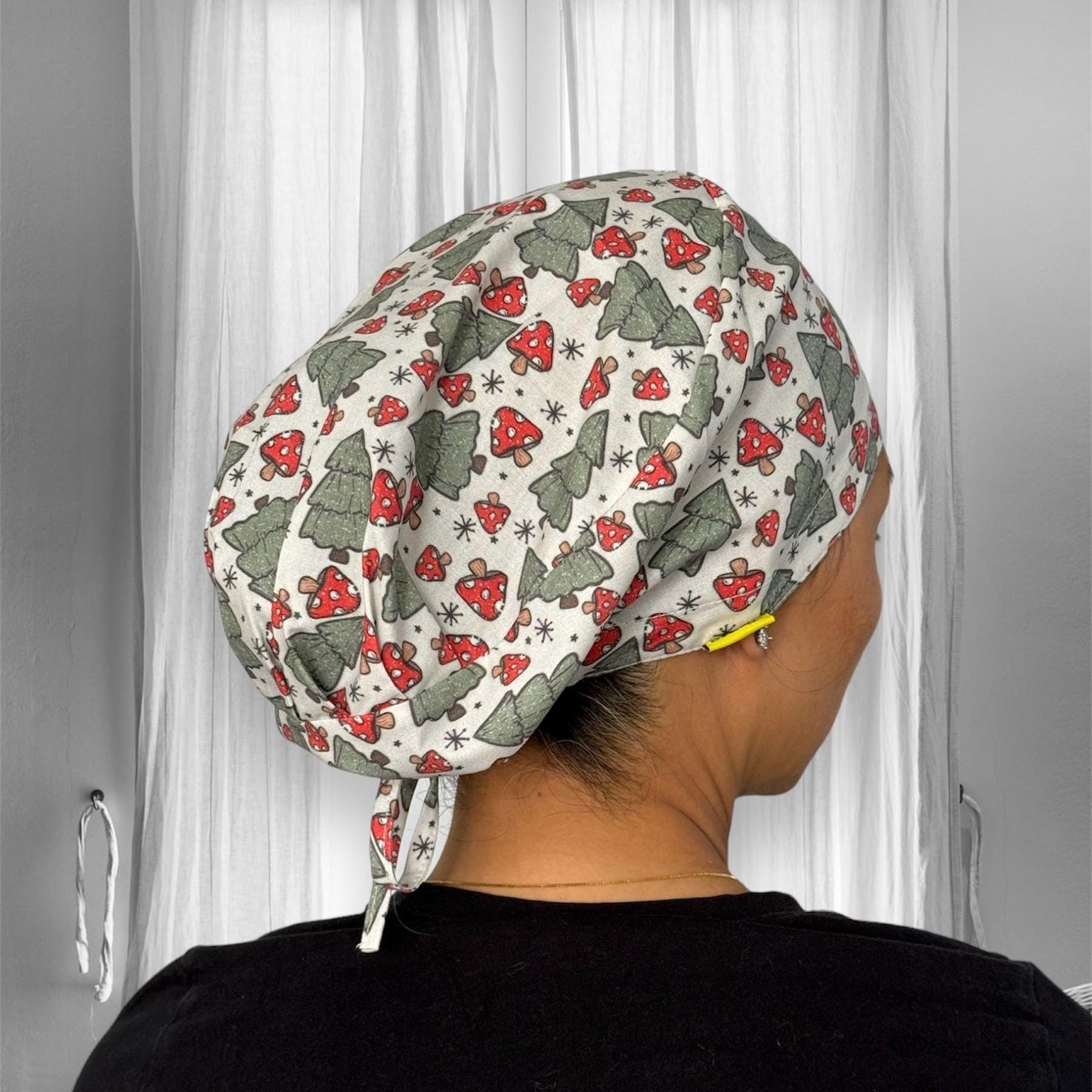 A scrub cap adorned with a festive pattern featuring green Christmas trees and red mushrooms, set against a light background. The cap covers the hair completely and is tied at the back.