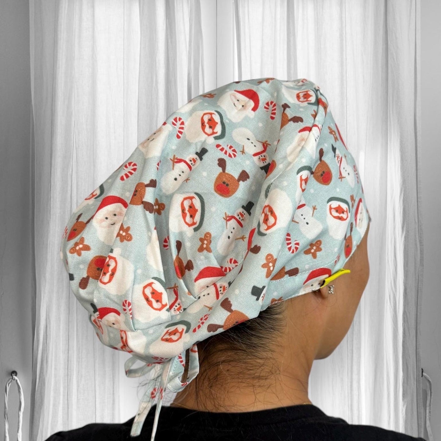 A scrub cap made from fabric decorated with festive designs, including images of Santa Claus, snowmen, candy canes, and gingerbread men. The cap is tied at the back and features a light blue background.