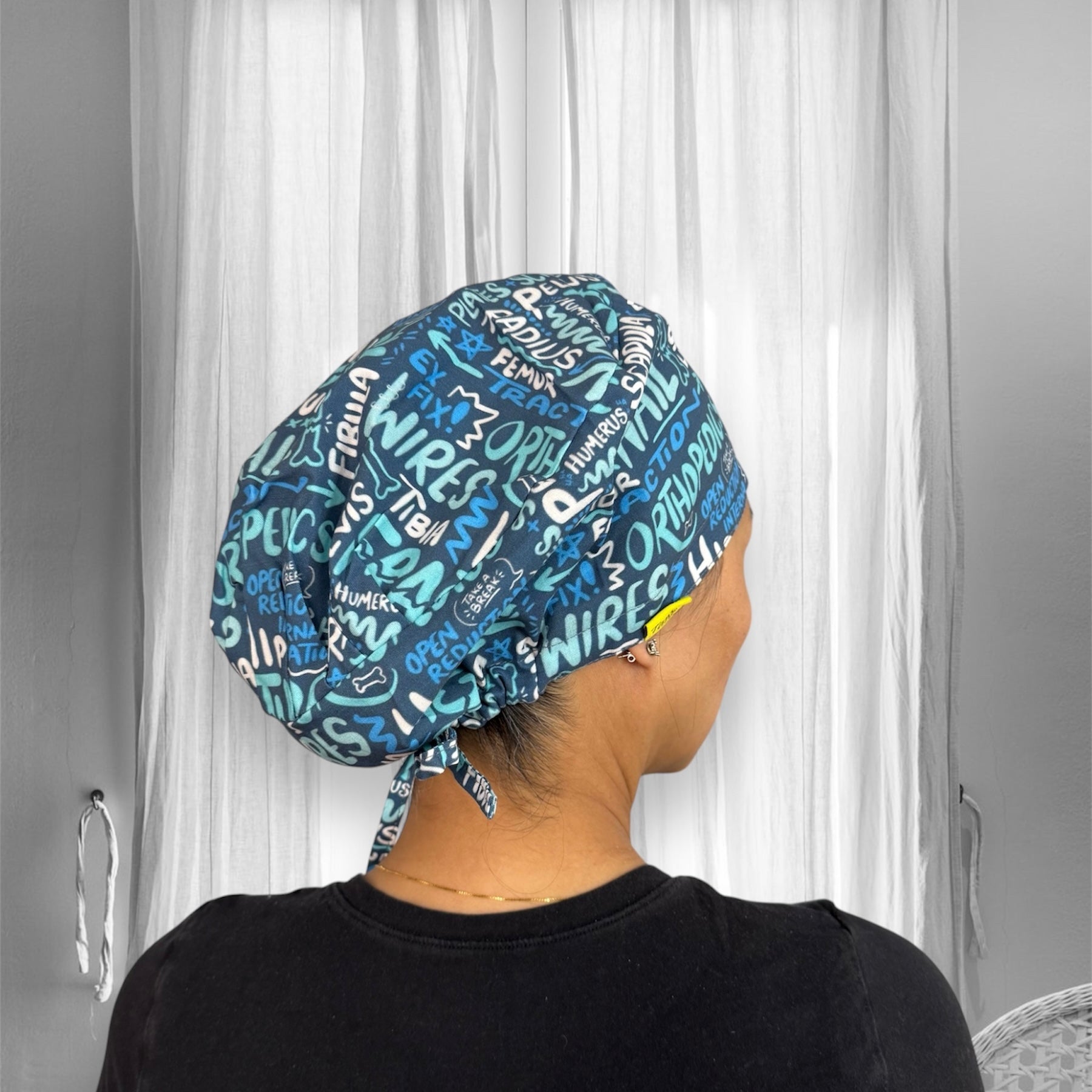 A person is wearing a blue scrub cap adorned with various medical terms and symbols in white and light blue text.