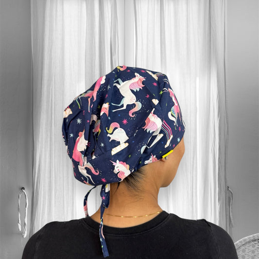 A person is wearing a scrub cap with a playful unicorn design. The cap features vibrant colors, including pink and white unicorns on a dark blue background, with stars and rainbow accents.