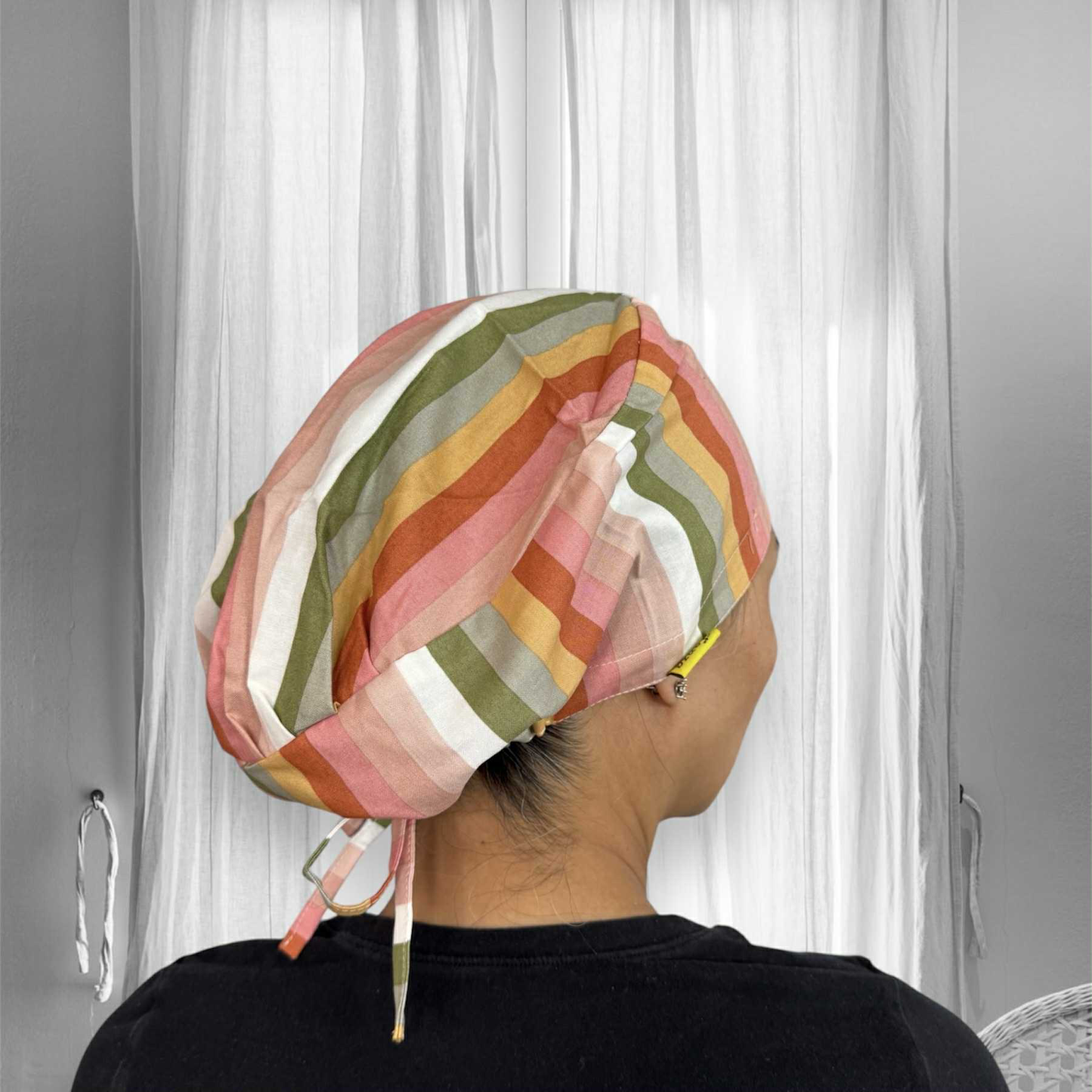 A person seen from behind, wearing a colorful scrub cap. The stripes on the head covering include shades of pink, orange, mustard yellow, olive green, and white.