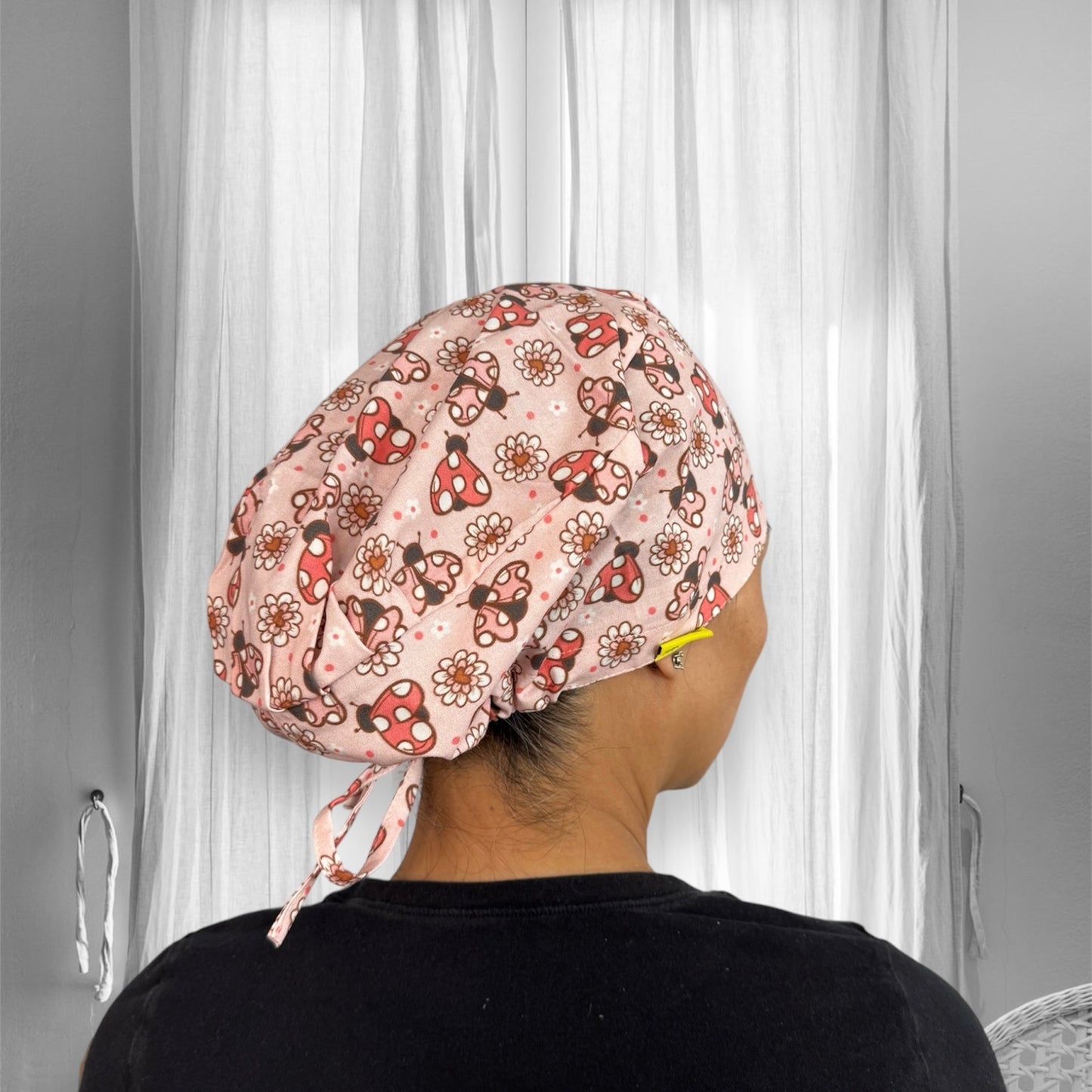 A person wearing a scrub cap featuring a playful pattern with red mushrooms, ladybugs, and white daisies on a light pink background.