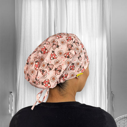 A person wearing a scrub cap featuring a playful pattern with red mushrooms, ladybugs, and white daisies on a light pink background.